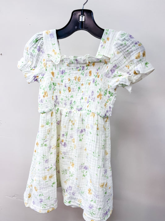 The Little Angel Dress by Tiny Tot Bundles is a short white floral dress with puffed sleeves, smocked bodice, and ruffled neckline, featuring pastel purple and yellow flowers. It hangs on a black hanger against a light-colored wall.