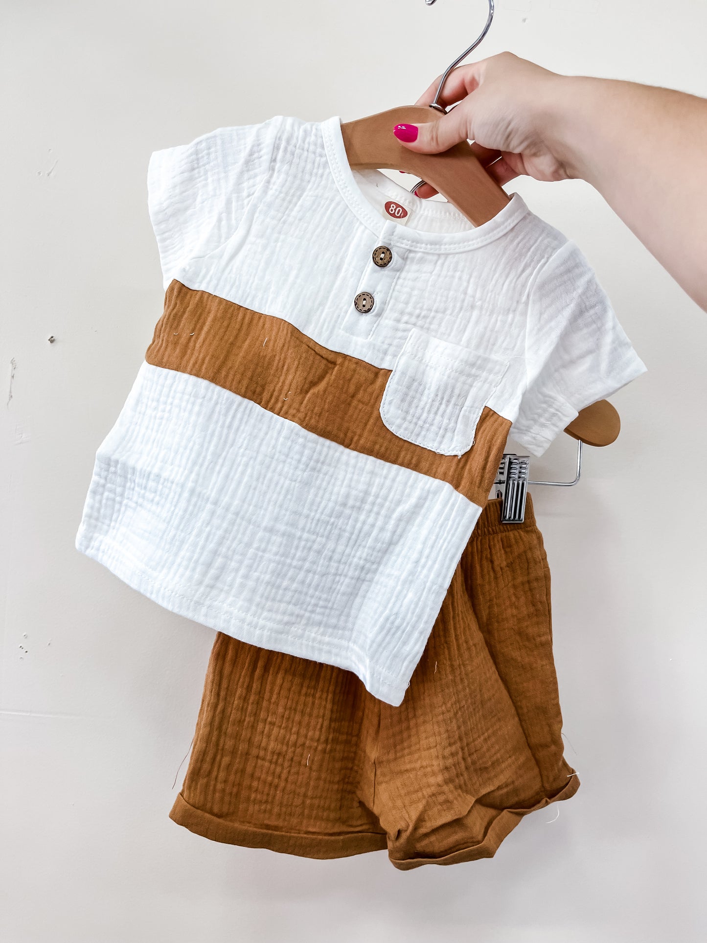 A hand holds Tiny Tot Bundles’ Linen Classic Set—a white short-sleeve top with brown stripes and buttons, plus matching brown shorts—displayed on a hanger against a plain white background.