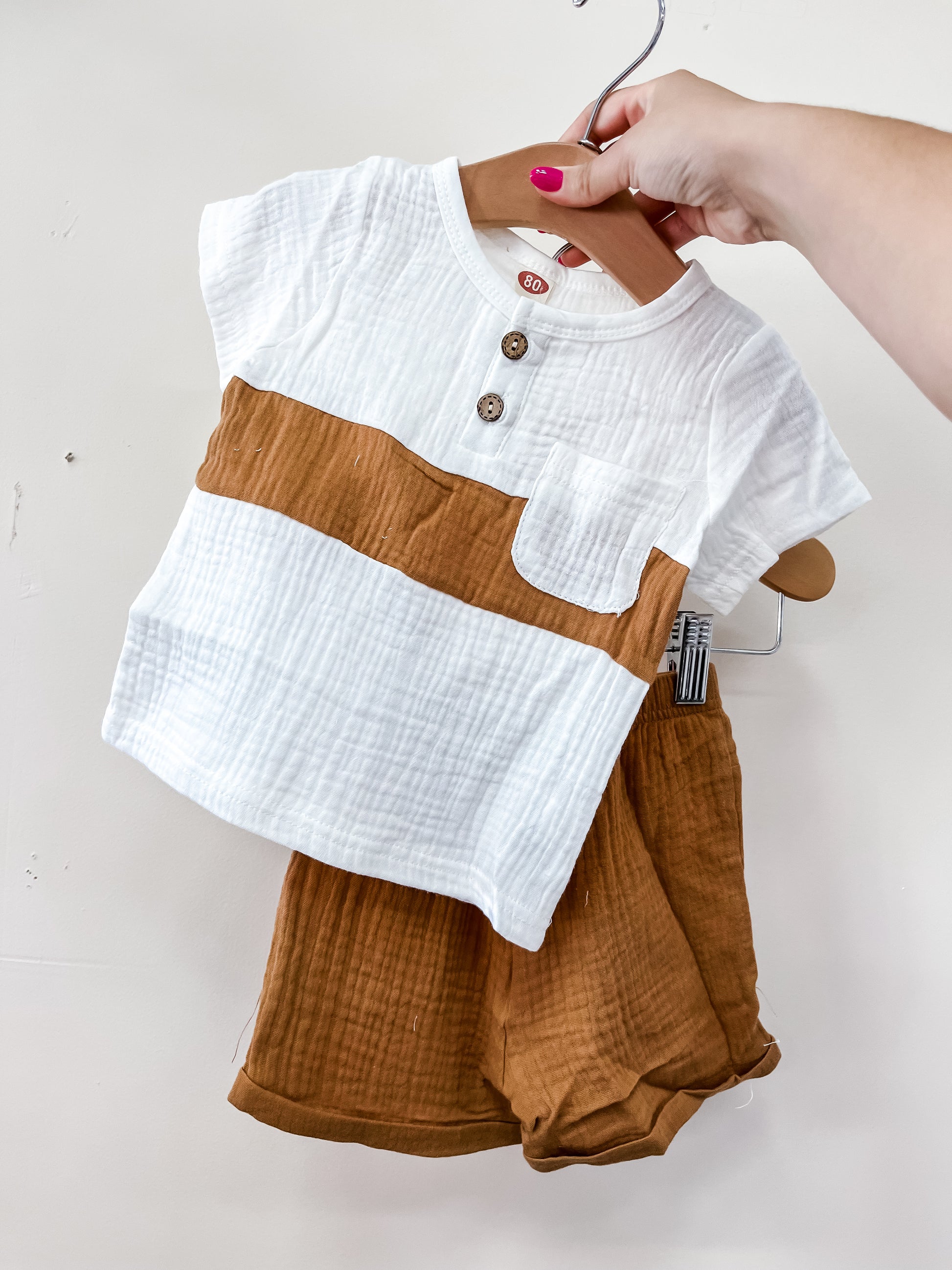 A hand holds Tiny Tot Bundles’ Linen Classic Set—a white short-sleeve top with brown stripes and buttons, plus matching brown shorts—displayed on a hanger against a plain white background.