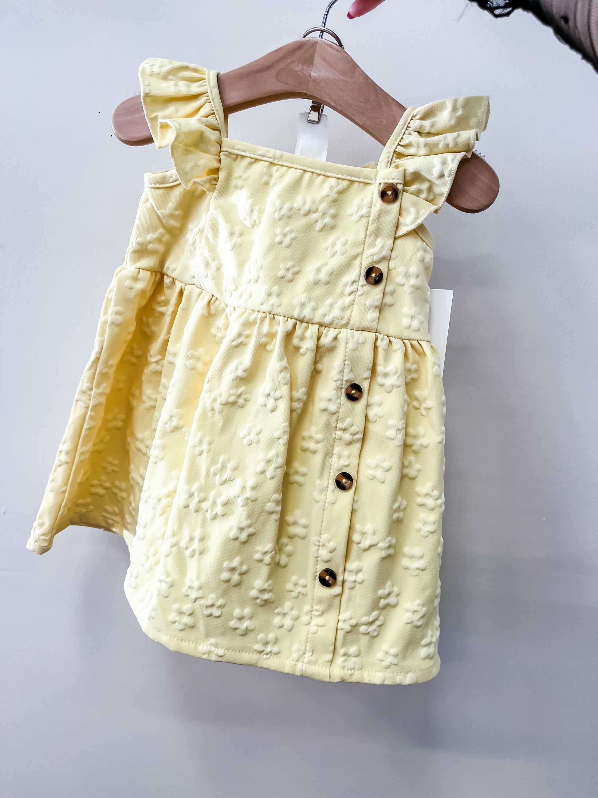 The Lemon Blossom Dress by Tiny Tot Bundles is a light yellow toddler dress with ruffled straps, a floral textured pattern, and brown front buttons, shown on a wooden hanger against a light background.