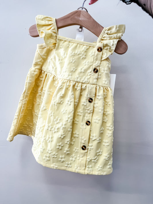 The Lemon Blossom Dress by Tiny Tot Bundles is a light yellow toddler dress with ruffled straps, a floral textured pattern, and brown front buttons, shown on a wooden hanger against a light background.