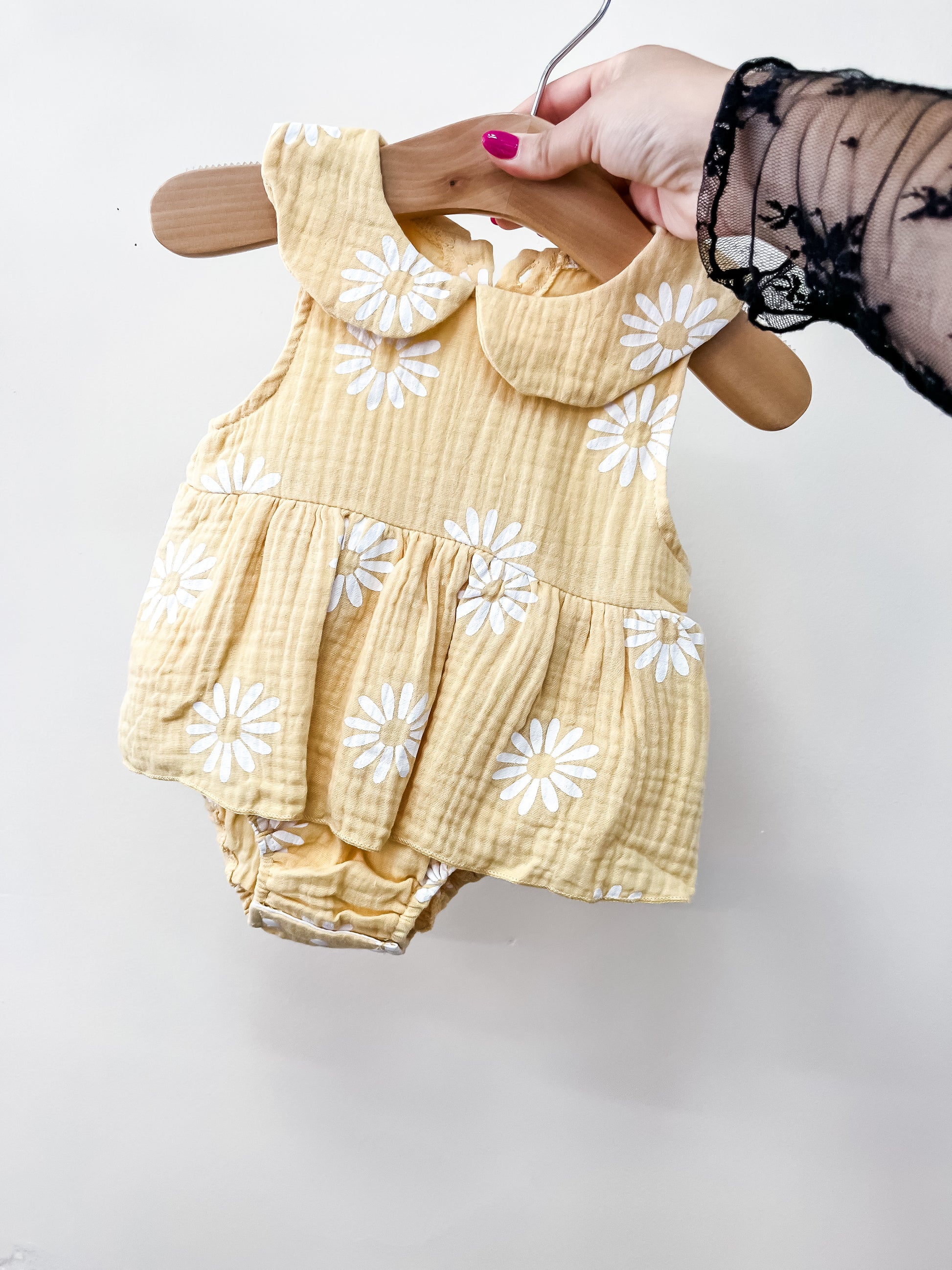 A hand holds the Tiny Tot Bundles Sunny Bloom Romper—a yellow baby outfit with white daisies, Peter Pan collar, and ruffled bottom—displayed on a wooden hanger against a plain white background.
