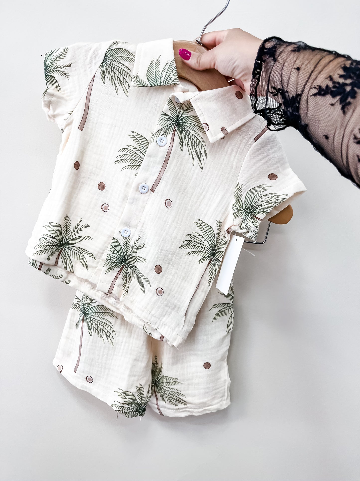 A hand holds up the Tiny Tot Bundles Palm Paradise Linen set—a matching two-piece baby outfit with palm tree and dot patterns, featuring a short-sleeved button-up shirt and shorts in light, textured fabric.