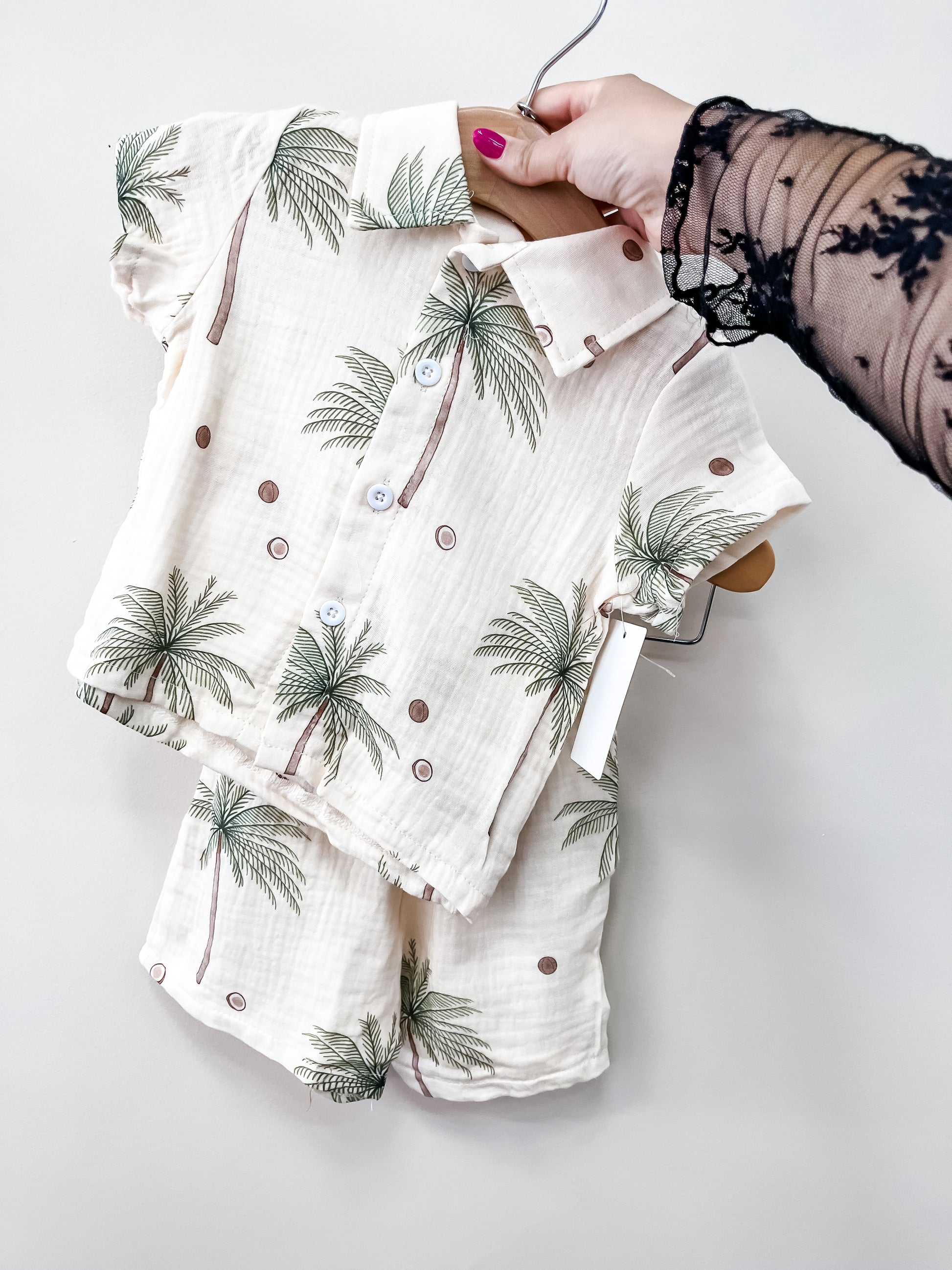 A hand holds up the Tiny Tot Bundles Palm Paradise Linen set—a matching two-piece baby outfit with palm tree and dot patterns, featuring a short-sleeved button-up shirt and shorts in light, textured fabric.