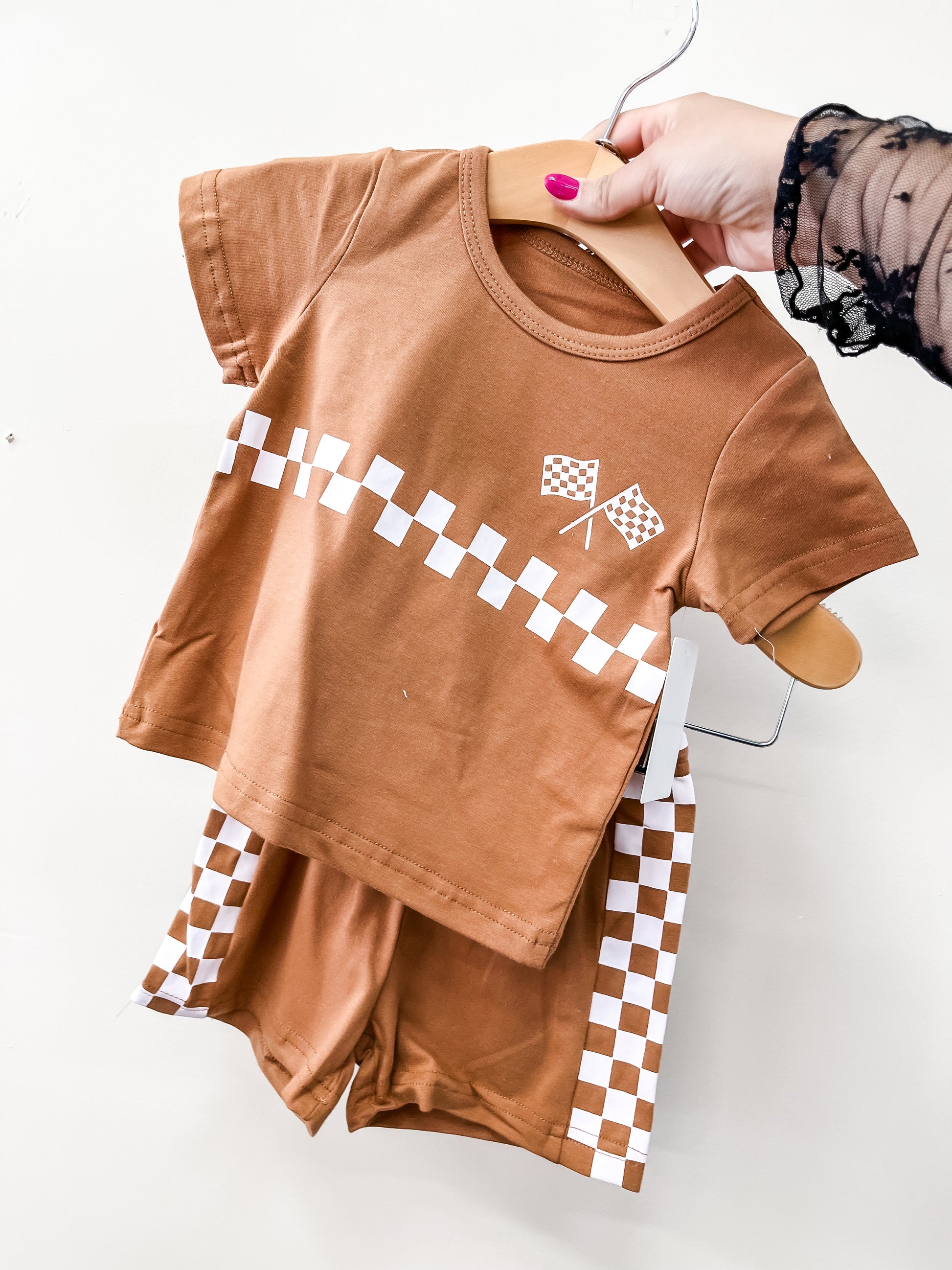A hand holds the Tiny Tot Bundles Rust Racer Check Set, a toddler outfit with a short-sleeve shirt and shorts featuring white and brown checkered patterns and two checkered flags on the shirt.