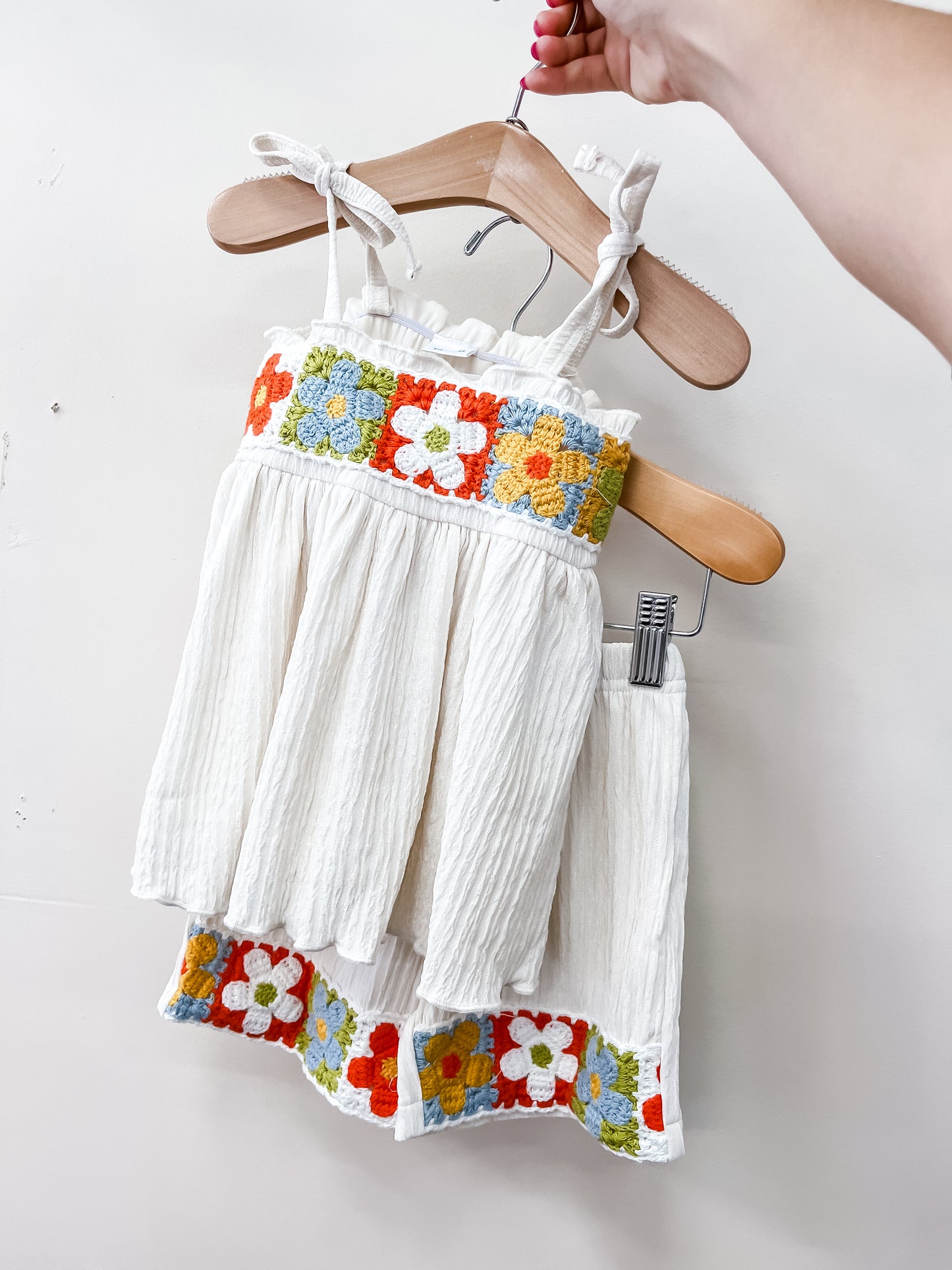 The Sunshine Crochet Set by Tiny Tot Bundles features a white sleeveless toddler dress and matching skirt with vibrant floral embroidery, shown on wooden hangers held by hand against a plain white background.