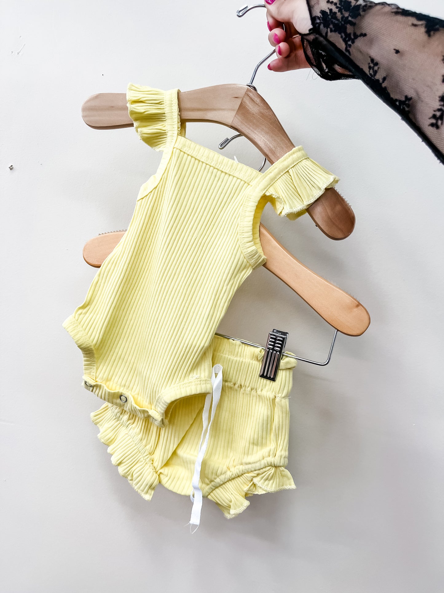 Someone holds up the Sunny Days Ribbed Set by Tiny Tot Bundles—a yellow ribbed baby romper with matching ruffled shorts—displayed against a plain white background.