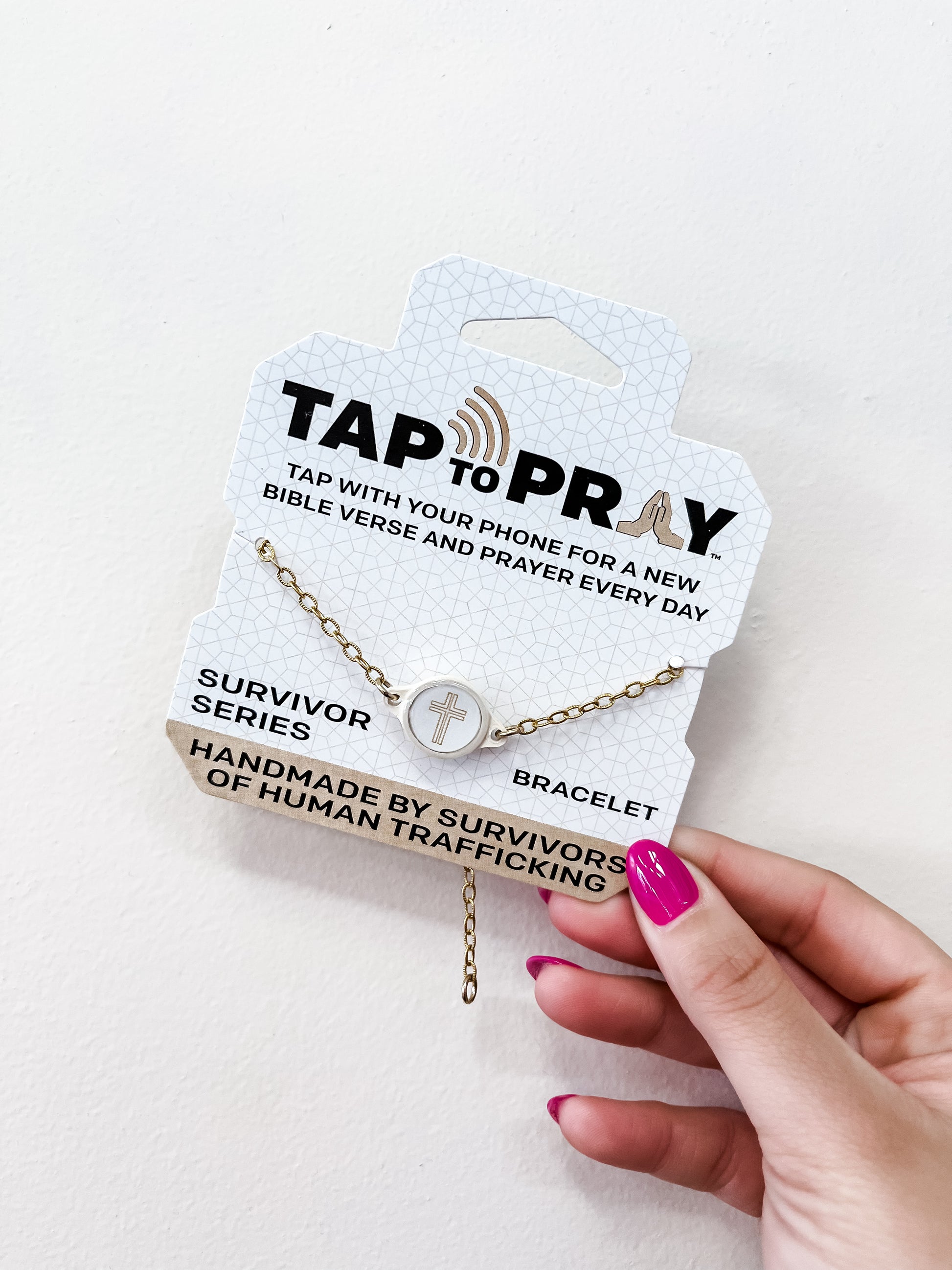 A hand with pink nails holds a versible Tap To Pray Bracelet - Gold Chain package, featuring a circular cross charm and noting it is handmade by survivors of human trafficking.