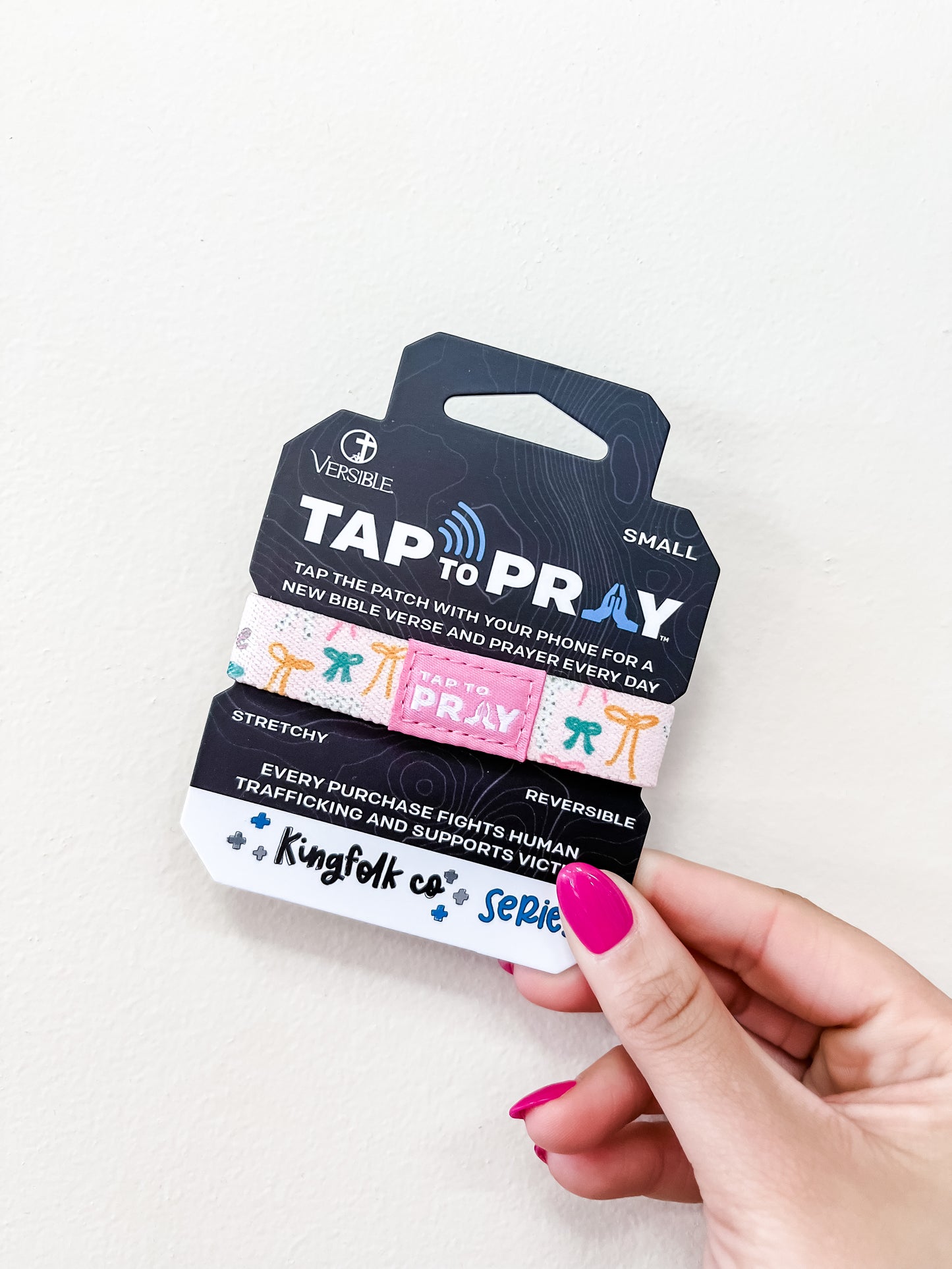 A hand with pink nail polish holds a packaged "Tap To Pray Bracelet - Fearfully & Wonderfully Made" by versible. The packaging says tap for daily Bible verses and that proceeds help fight human trafficking.