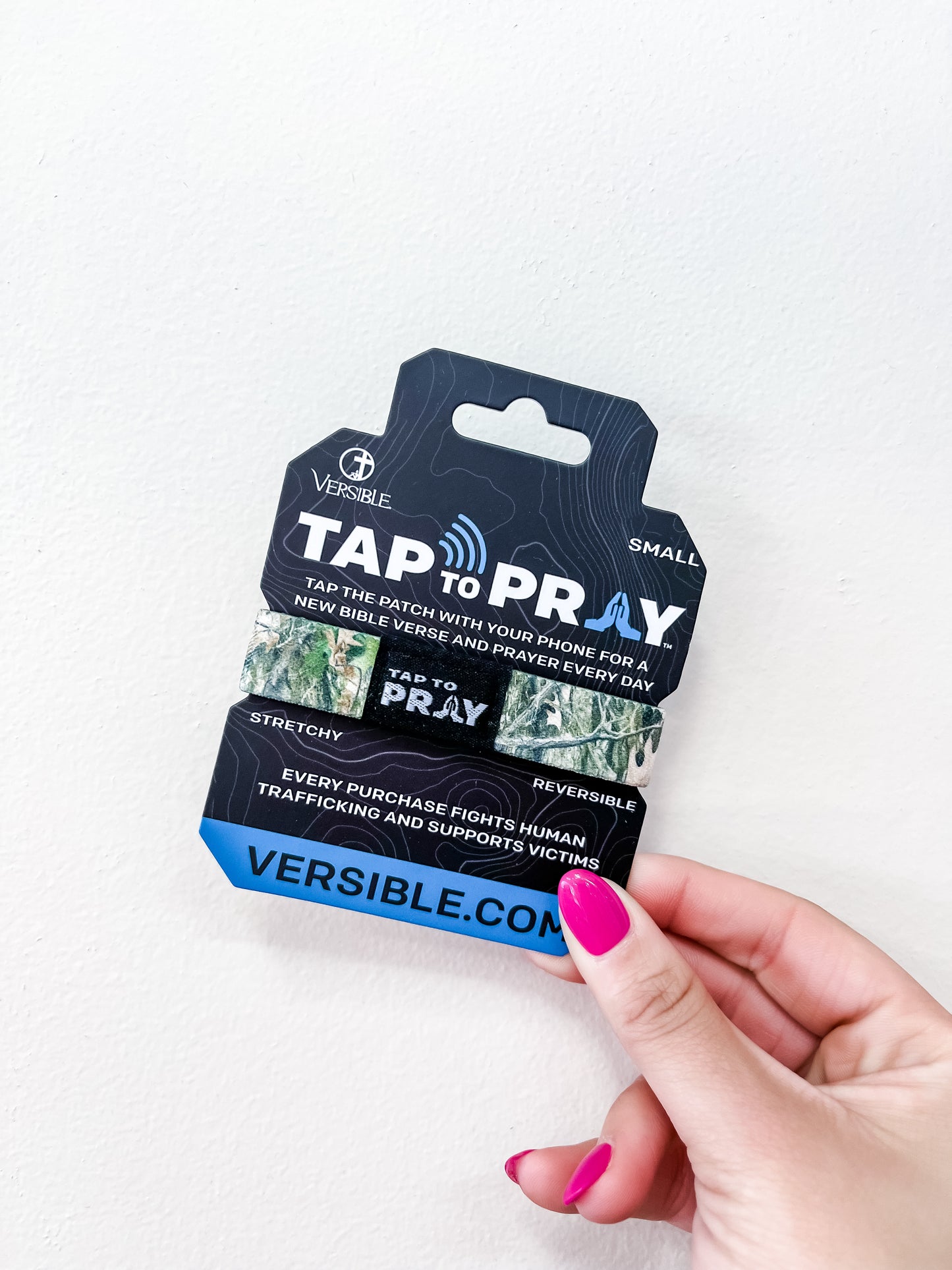 A hand with pink nail polish holds a versible Tap To Pray Bracelet - Forest Camo package, featuring a stretchy, reversible band. The packaging highlights Bible verses, prayer, and support for human trafficking victims.