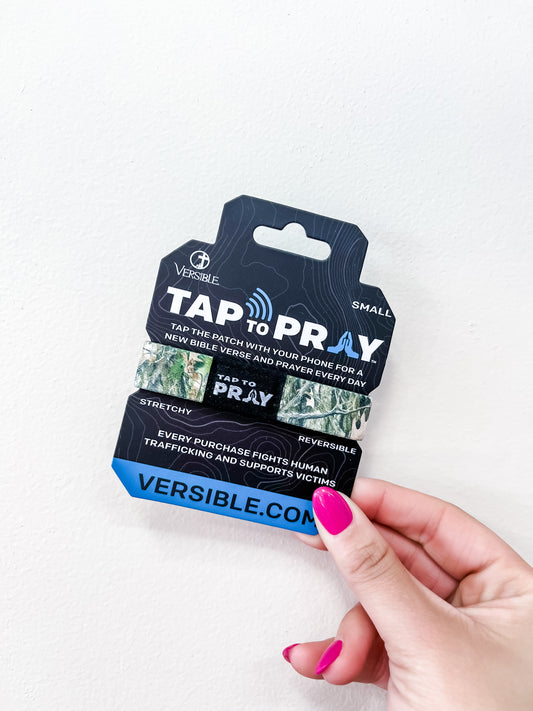 A hand with pink nail polish holds a versible Tap To Pray Bracelet - Forest Camo package, featuring a stretchy, reversible band. The packaging highlights Bible verses, prayer, and support for human trafficking victims.