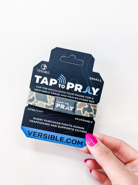 A hand with pink nail polish holds a versible Tap To Pray Bracelet - Sahara Camo package, supporting human trafficking victims and featuring the website VERSIBLE.COM.