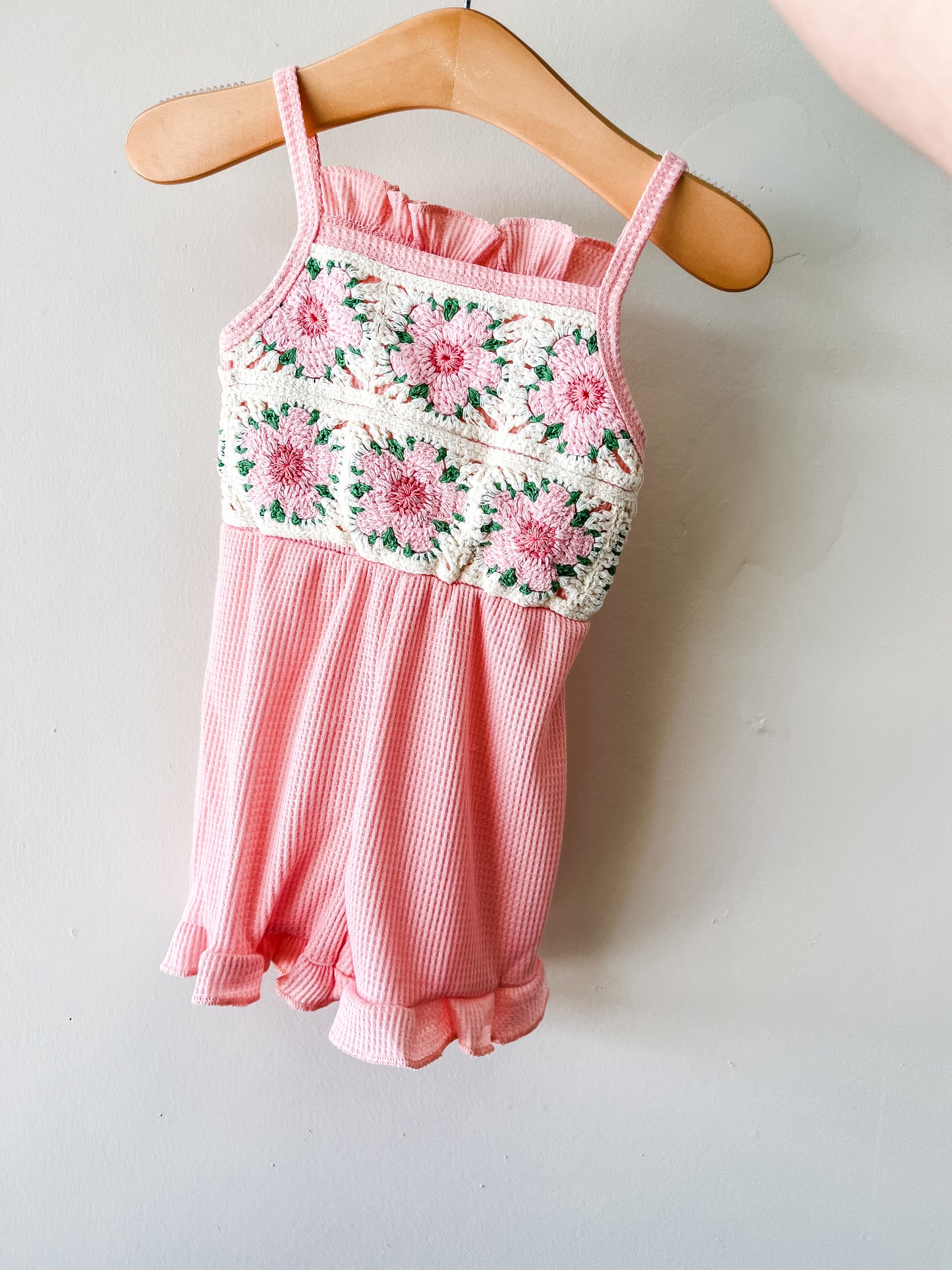 The Pink Crochet Romper by Tiny Tot Bundles is a pink sleeveless child’s outfit with a crocheted top featuring pink and green flowers and ruffled-hem shorts, displayed on a wooden hanger.