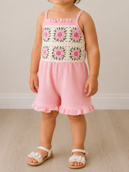 A young child wears the Tiny Tot Bundles Pink Crochet Romper indoors on a wooden floor, paired with white sandals featuring bow details.