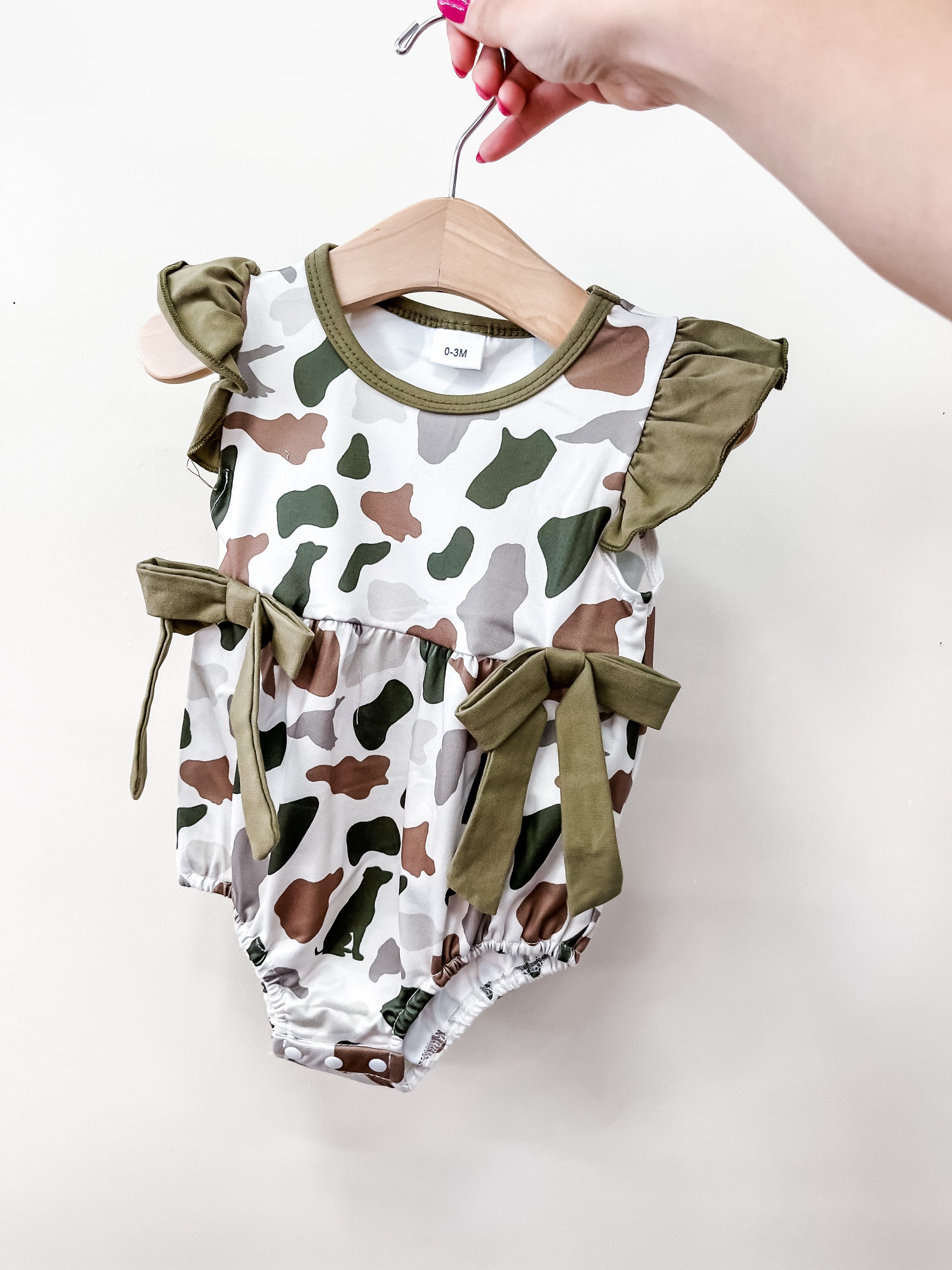 A hand holds the Tiny Tot Bundles Green Camo Bow Romper, featuring a green, brown, and white camouflage pattern with ruffled sleeves and waist bows, against a plain white background.