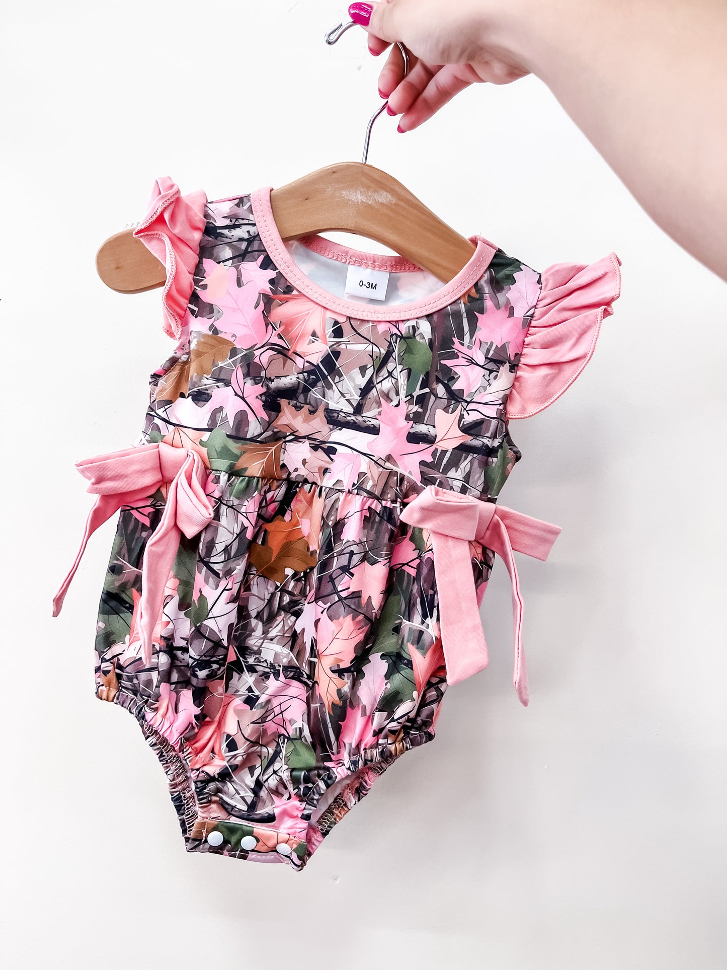 A hand displays the Tiny Tot Bundles Pink Camo Bow Romper on a wooden hanger, featuring a pink camo pattern, ruffled sleeves, and pink bow accents on each side against a plain white background.
