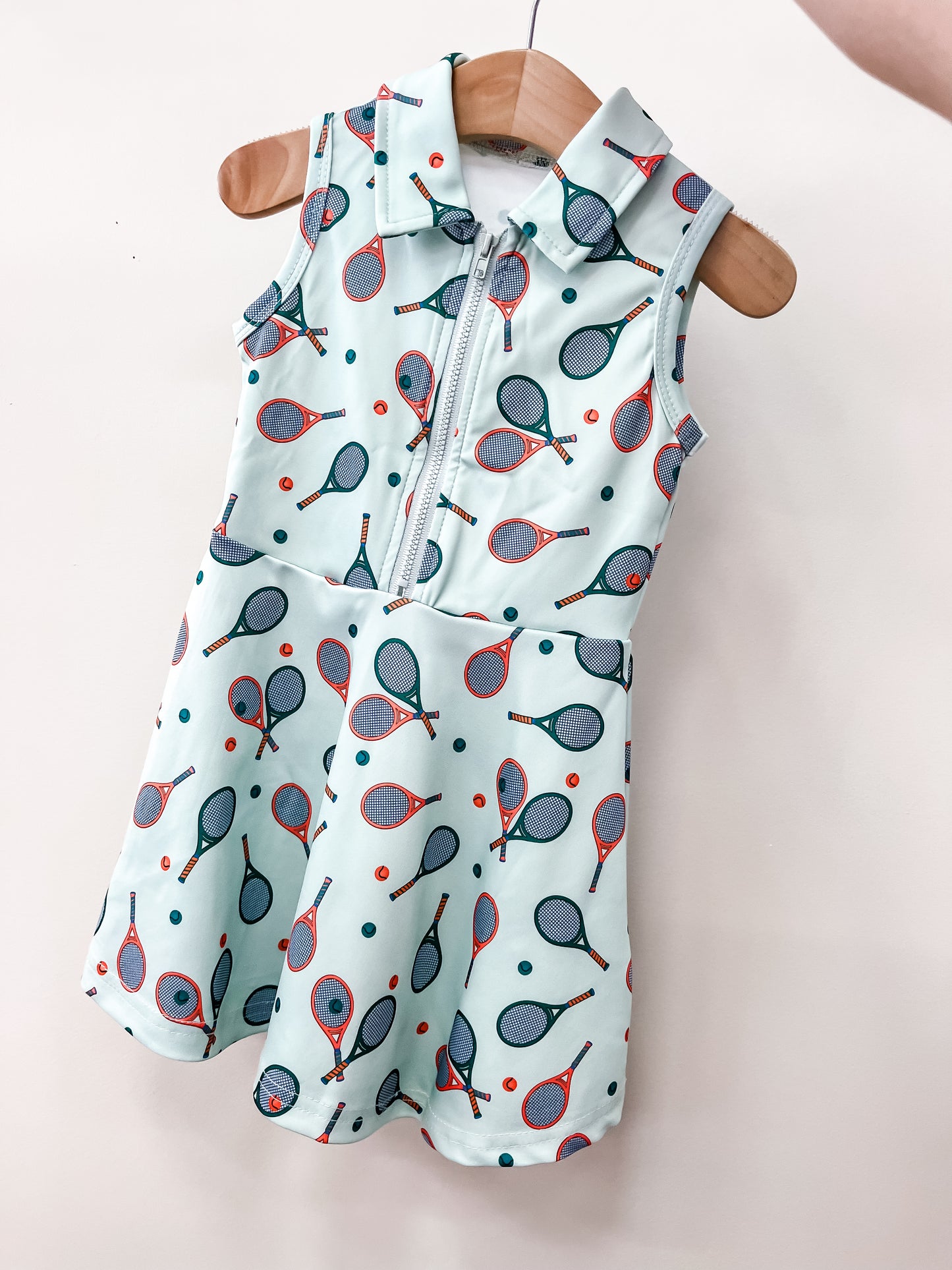 The Tiny Tot Bundles Green Quarter Zip Tennis Dress for children features a sleeveless design with a tennis racket and ball pattern, displayed on a wooden hanger against a white background.