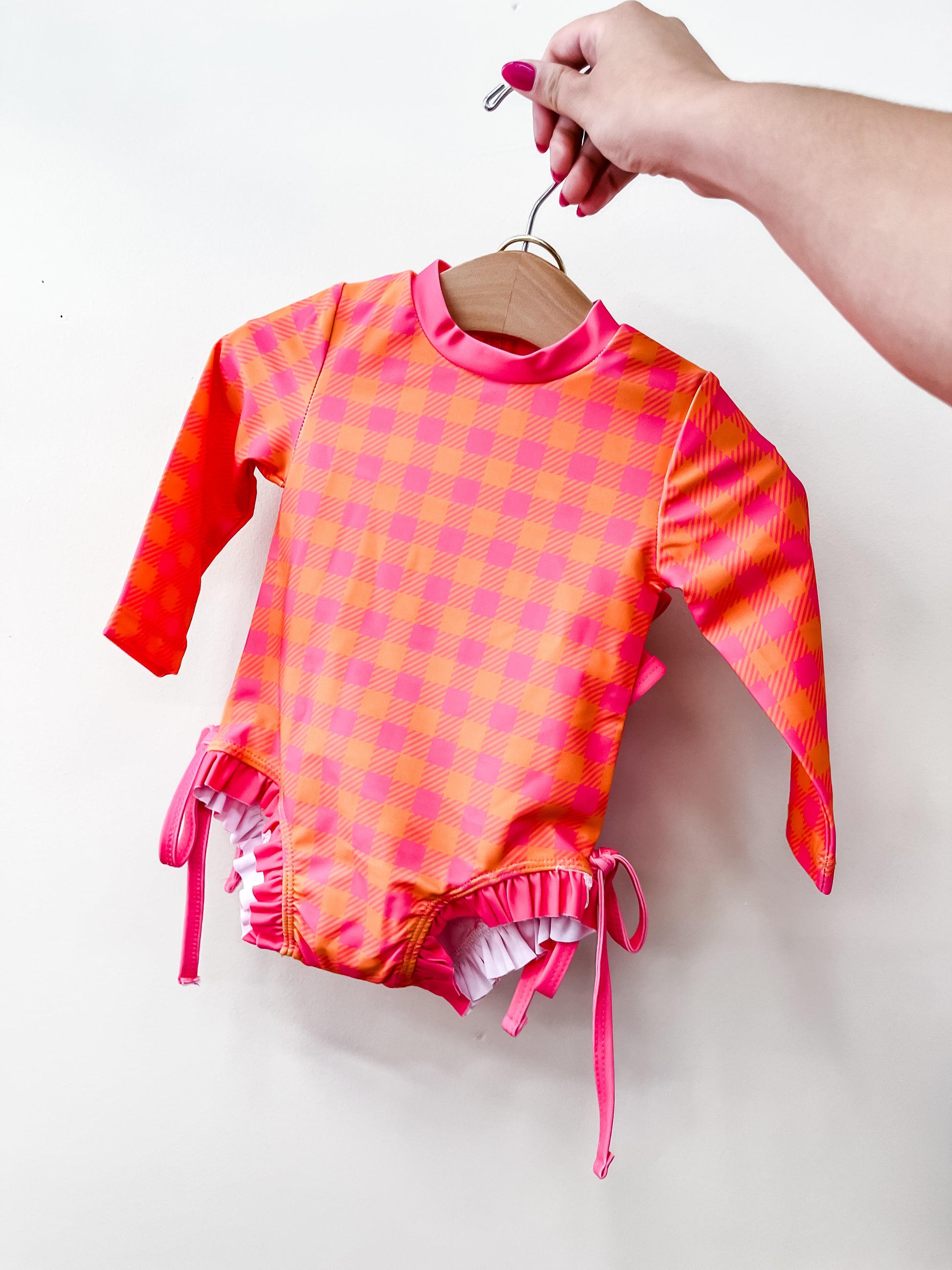 A hand holds Tiny Tot Bundles’ Sunset Splash Check Swimsuit, a long-sleeve toddler suit with pink and orange checkers, side ties, and ruffles, displayed on a hanger against a plain white background.