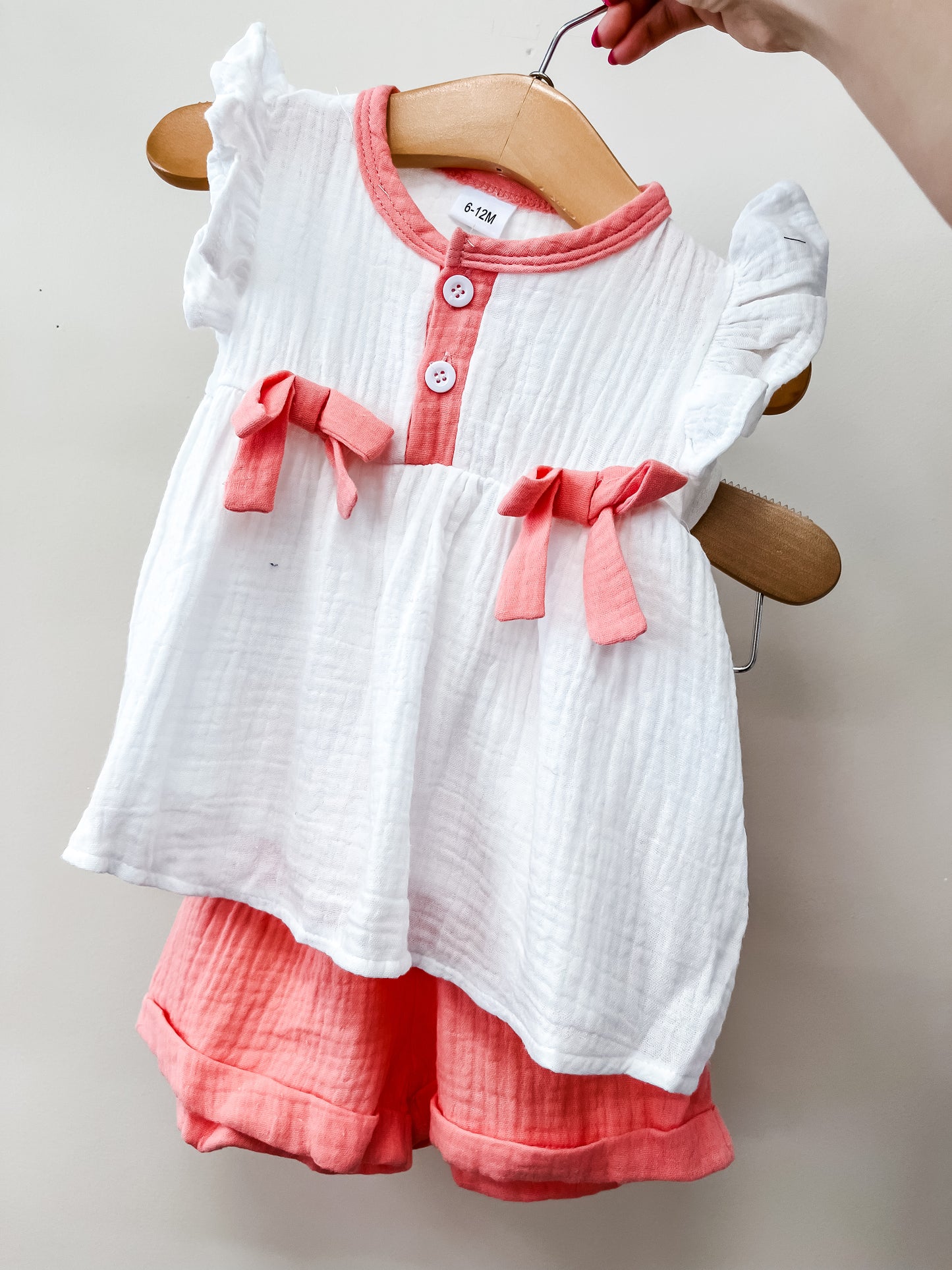 A hand displays the Peaches & Cream Short Set by Tiny Tot Bundles, featuring a white short-sleeve top with pink trim, buttons, bow details, and matching pink cuffed shorts.