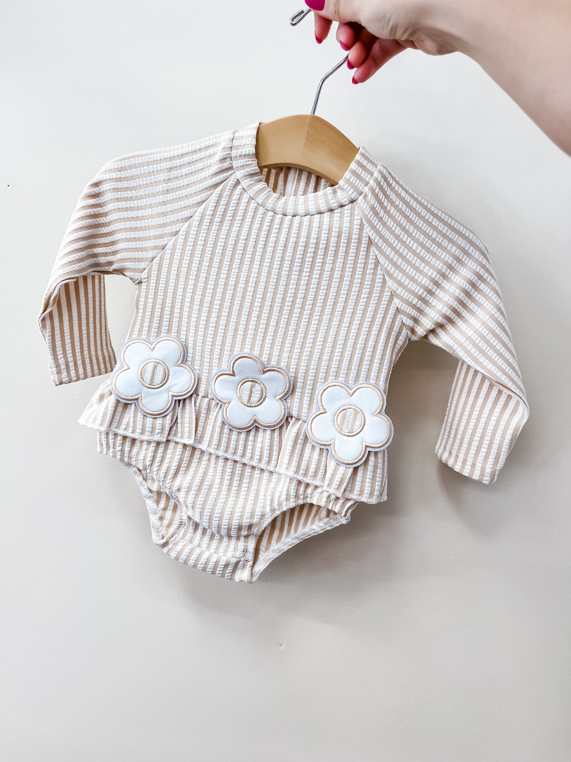 A hand holds a hanger displaying the Tiny Tot Bundles Petal Lane Swimsuit, featuring beige and white stripes, long sleeves, and three white flower appliqués on the ruffled waist.