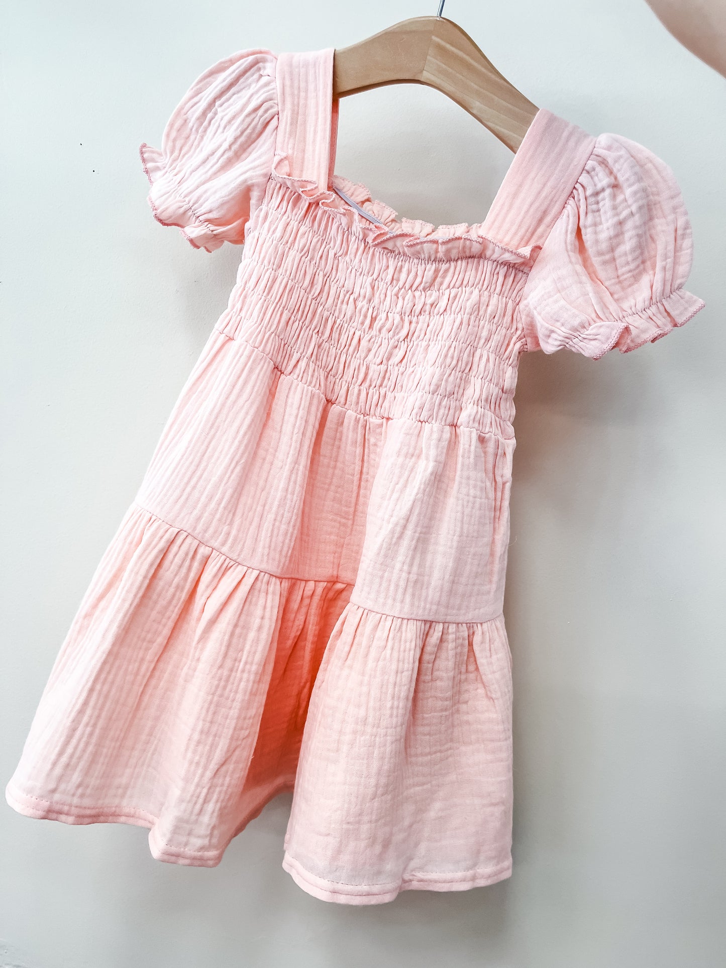The Blush Breeze Smocked Dress by Tiny Tot Bundles is a light pink, short-sleeved baby dress with puffed sleeves, ruffled trim, and a smocked bodice, shown on a wooden hanger against a plain white background.