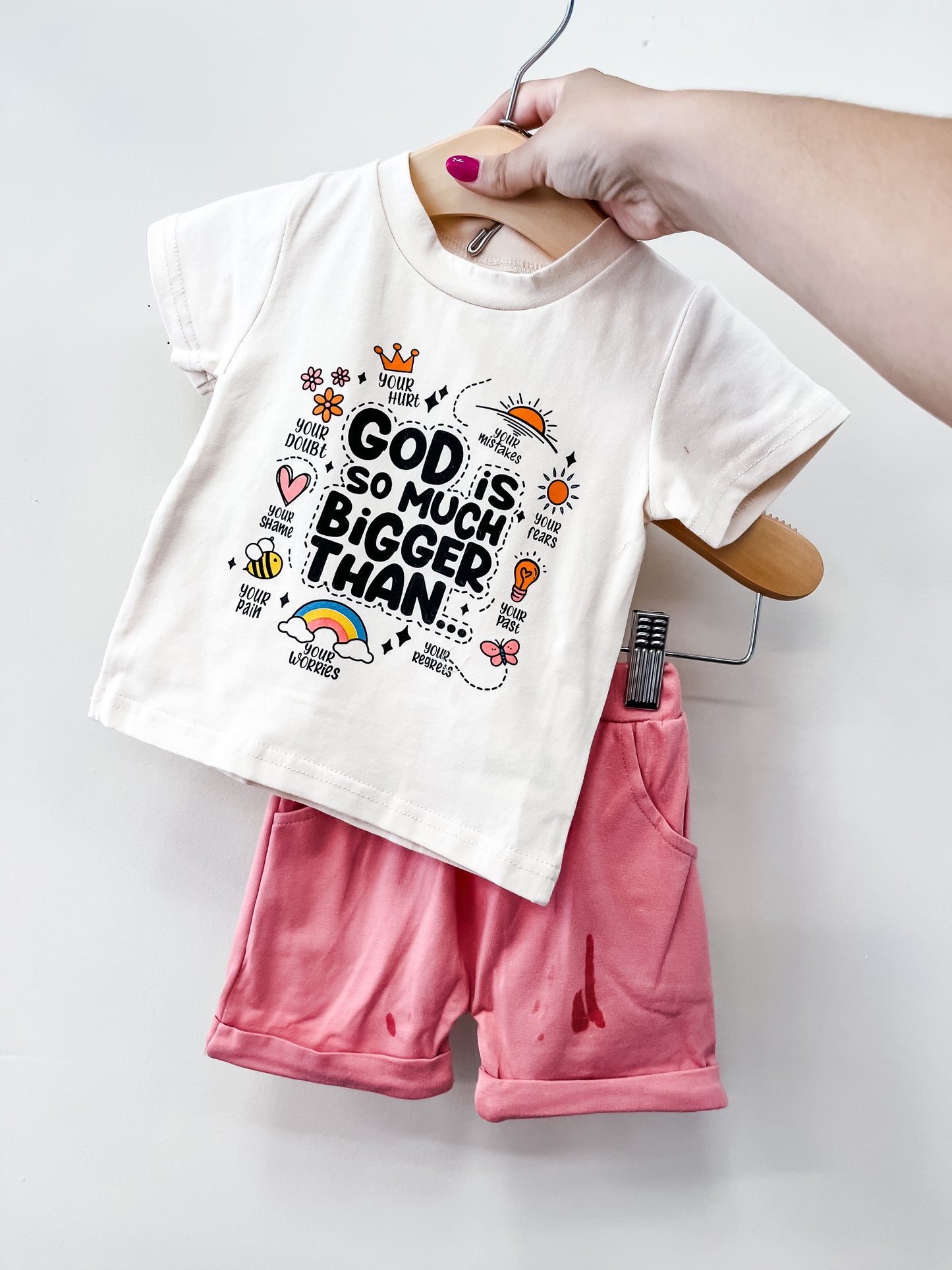 A hand holds the Tiny Tot Bundles God Is Bigger Short Set: a white tee with "God is so much bigger than" and colorful doodles, plus rolled-cuff pink shorts, displayed against a plain light background.