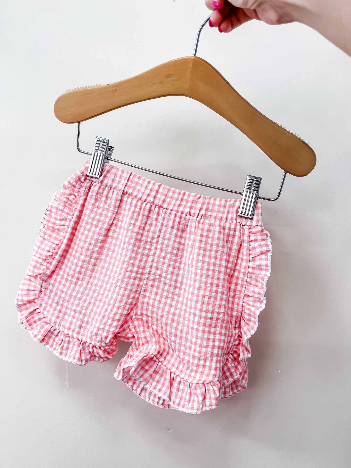 A hand holds a wooden hanger with Tiny Tot Bundles’ Picnic Day Gingham Shorts—pink and white ruffled gingham—shown against a plain white background.