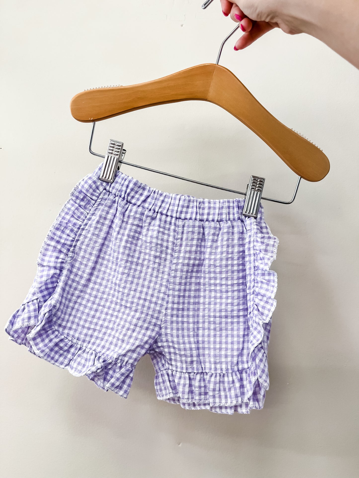 A hand holds a wooden hanger displaying the Picnic Day Gingham Shorts by Tiny Tot Bundles in light purple and white against a plain white background.