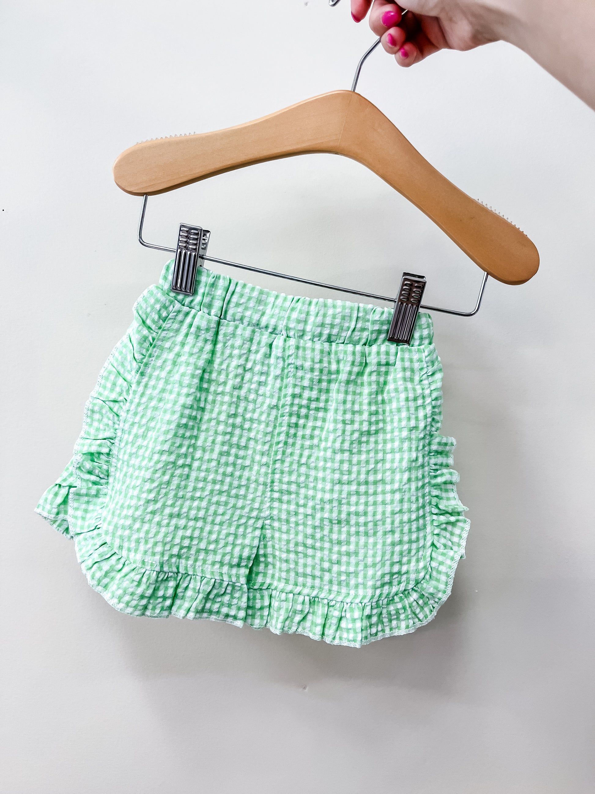 A hand holds Tiny Tot Bundles’ Picnic Day Gingham Shorts—light green and white with ruffles—displayed on a wooden hanger against a plain white background.