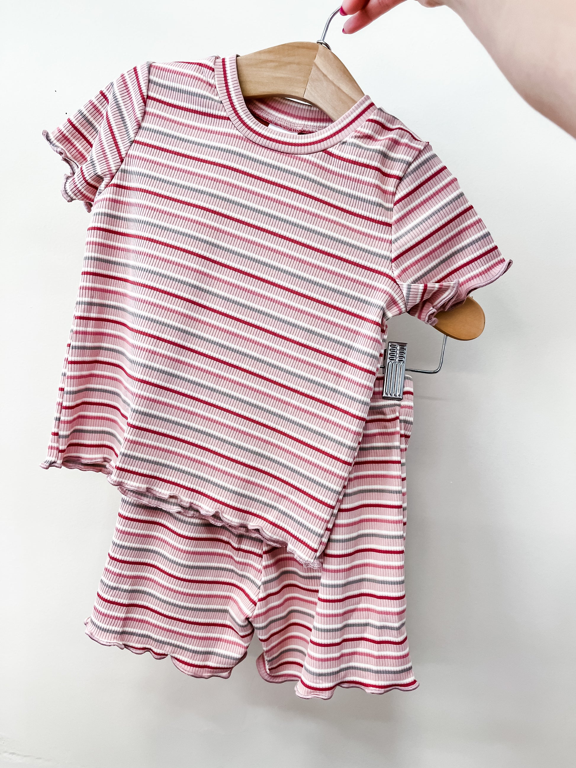 A hand holds a Tiny Tot Bundles Rosy Stripe Snuggle Set—a matching children's outfit with a pink and white striped, short-sleeved top and ruffled shorts—on a wooden hanger against a plain white background.