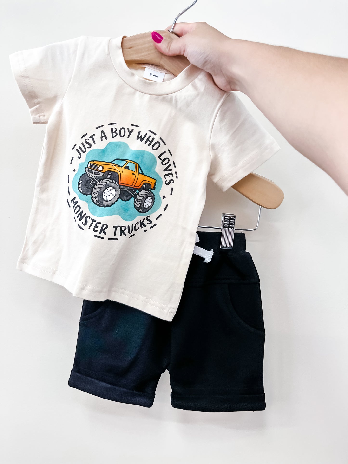 A hand holds the Tiny Tot Bundles Monster Truck Mania Set: a beige T-shirt with a monster truck graphic and "Just a boy who loves monster trucks," paired with black shorts.