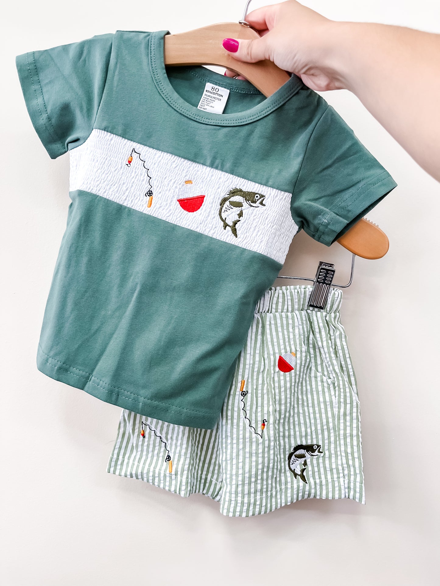 A hand holds the Little Angler Short Set by Tiny Tot Bundles: a green toddler t-shirt with fishing-themed embroidery and matching green-and-white striped shorts, both displayed on wooden hangers against a plain background.