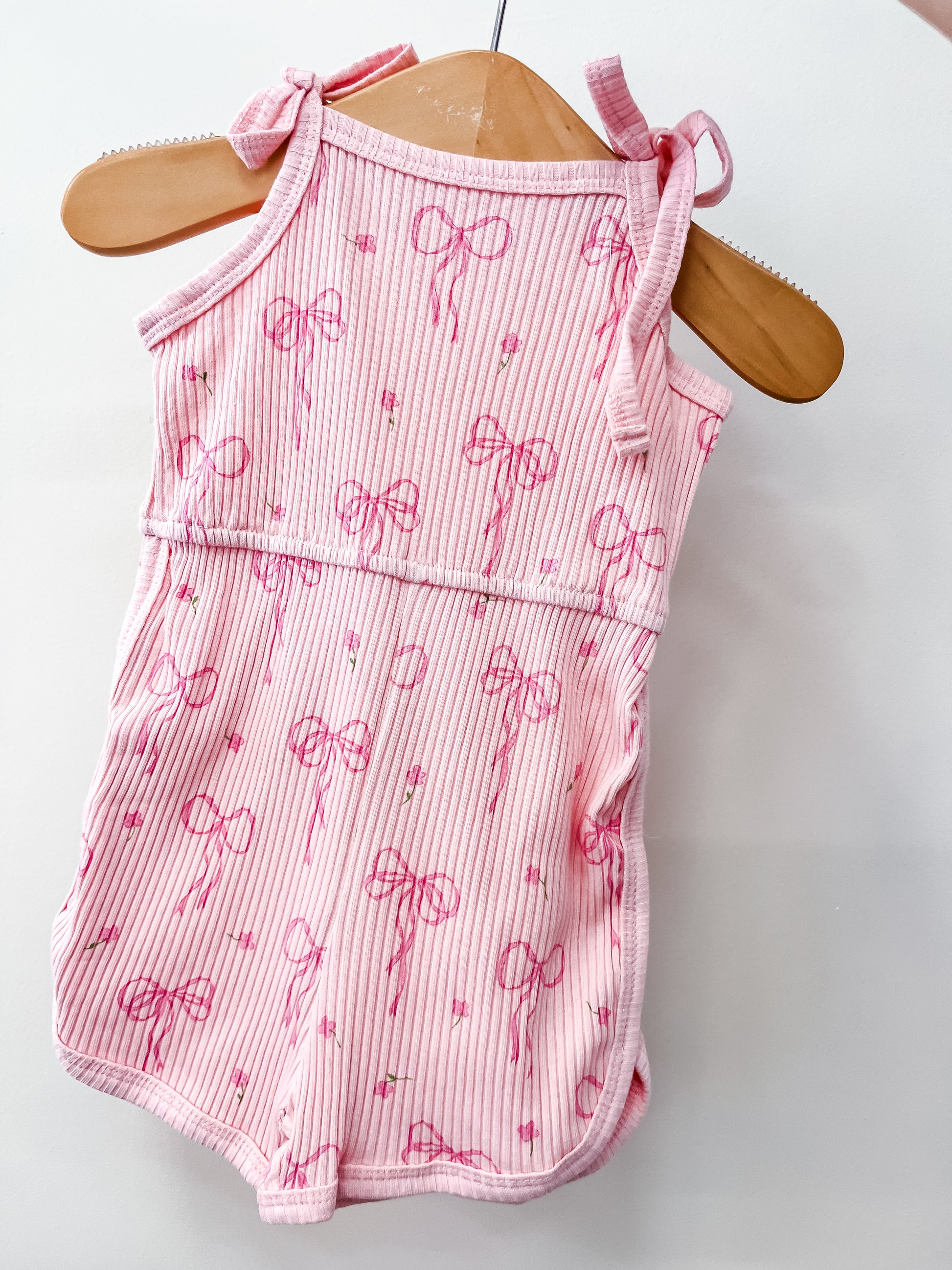 The Tiny Tot Bundles Pink Ribbon Tie Romper features shoulder ties and a pattern of darker pink bows with small stars, displayed on a wooden hanger against a white background.