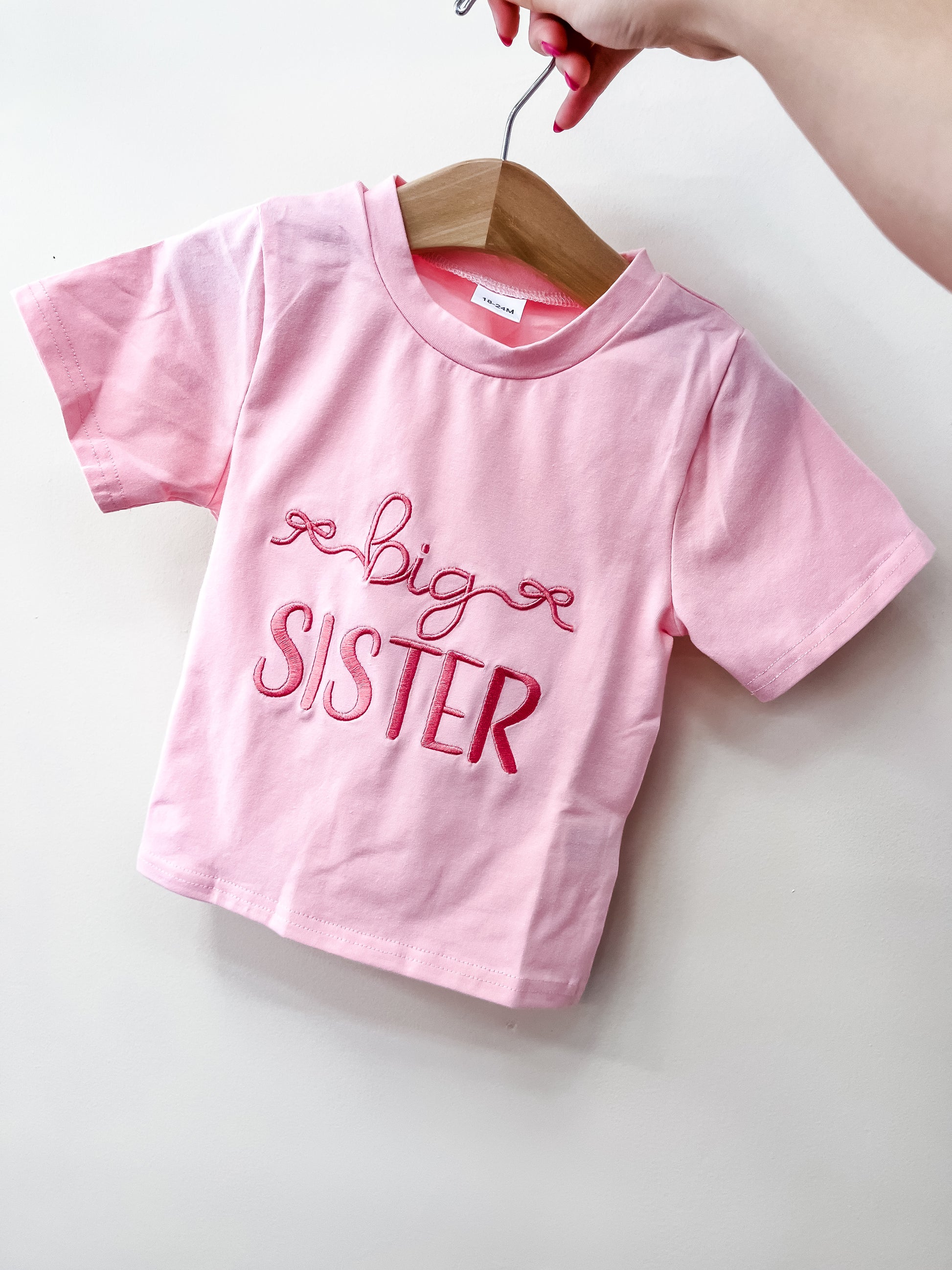 A hand holds a hanger with a pink Tiny Tot Bundles Big Sister Bow T-Shirt featuring "big SISTER" embroidered in red and pink script on the front.