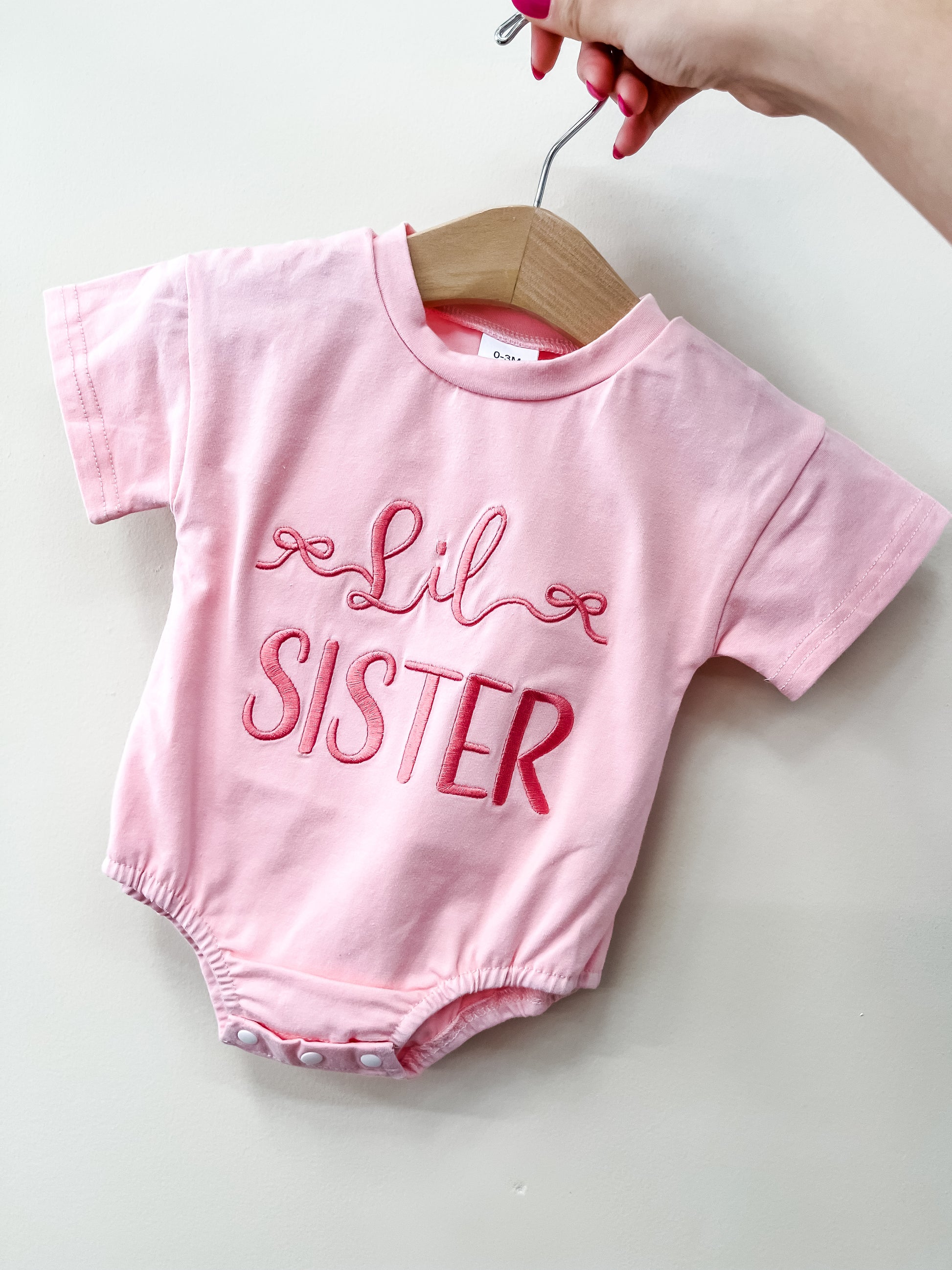 A hand holds a wooden hanger with the Tiny Tot Bundles Lil Sister Pink Bubble, a pink baby onesie featuring "Lil Sister" in bold decorative lettering on the front.