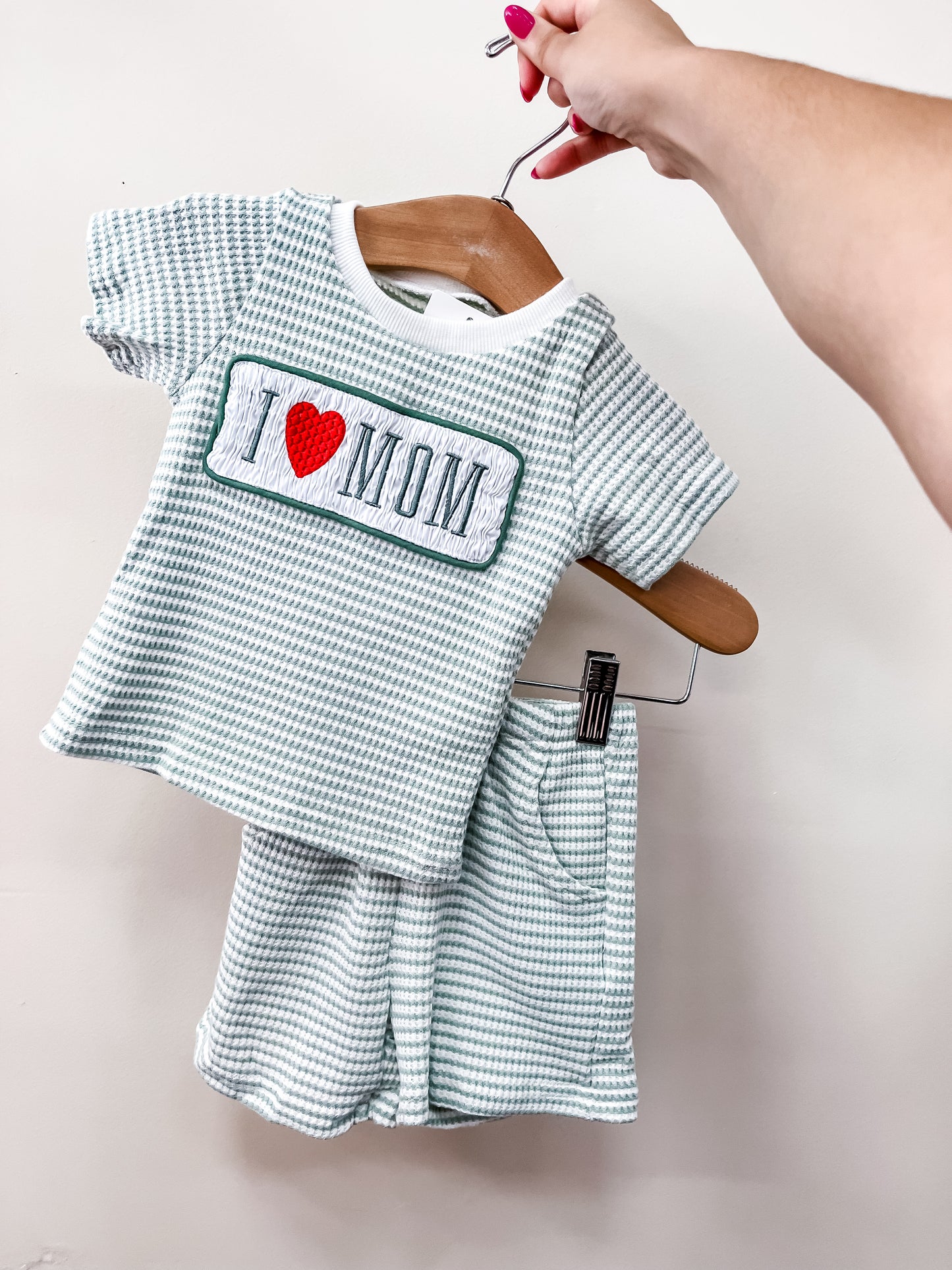 A hand holds the Tiny Tot Bundles Love Mom Stripe Set, a toddler’s outfit with a green and white checkered top featuring "I ♥ MOM" and matching shorts with an elastic waistband and pocket, against a plain background.