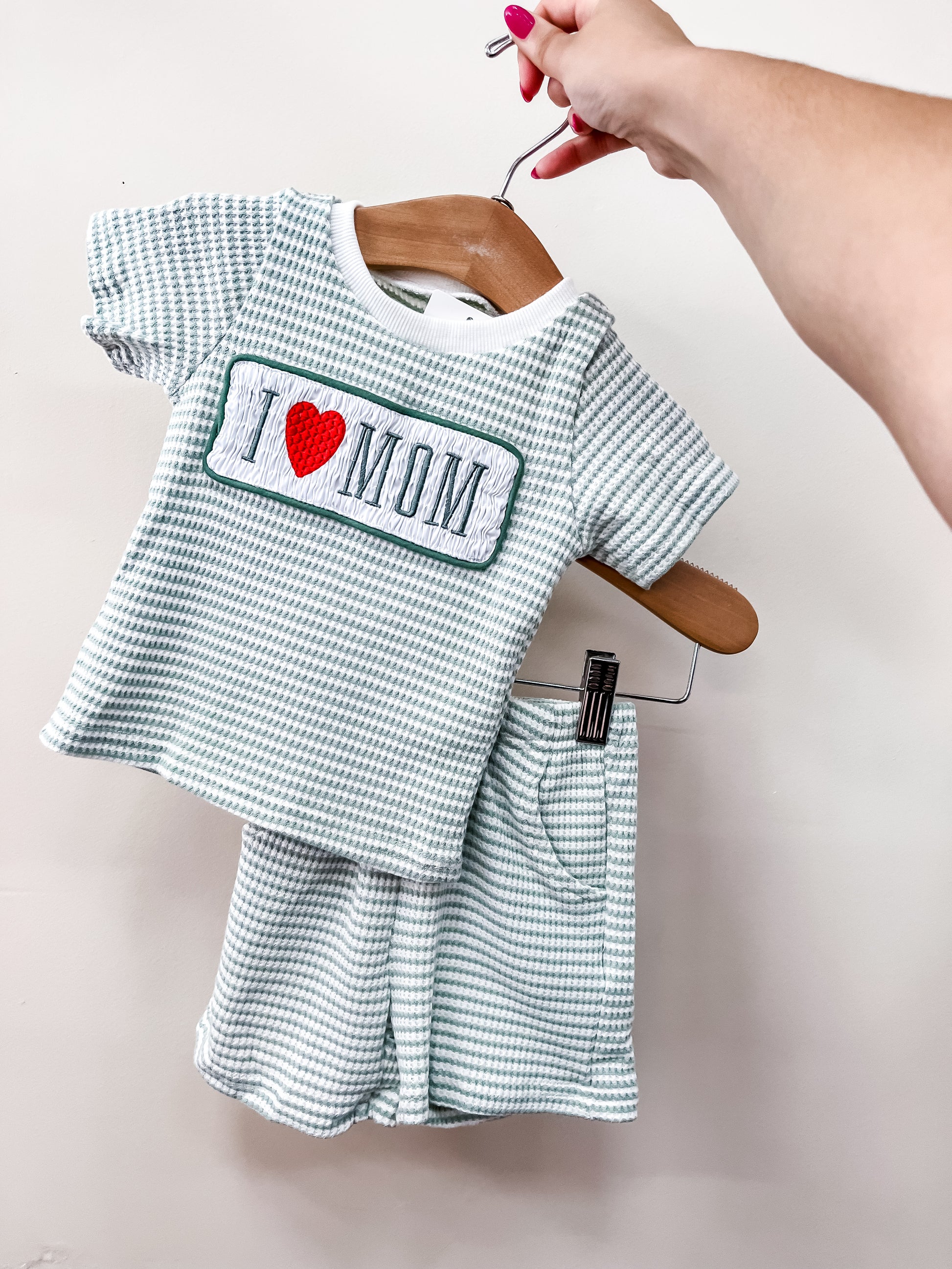 A hand holds the Tiny Tot Bundles Love Mom Stripe Set, a toddler’s outfit with a green and white checkered top featuring "I ♥ MOM" and matching shorts with an elastic waistband and pocket, against a plain background.