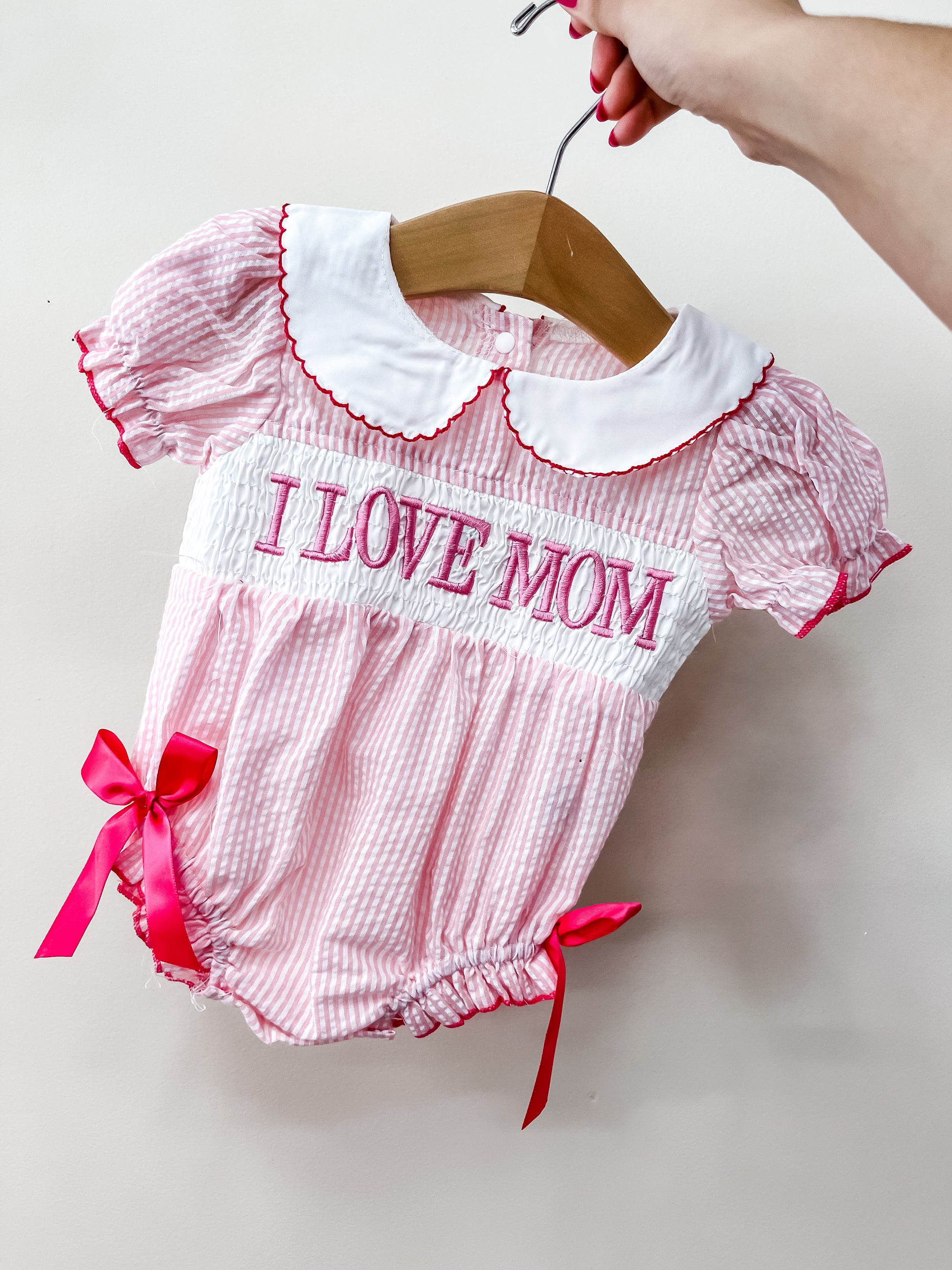 A hand holds the Tiny Tot Bundles "I Love Mom Bubble," a pink and white striped baby romper with a white Peter Pan collar, red trim, bows, and “I LOVE MOM” embroidered in large pink letters across the chest.