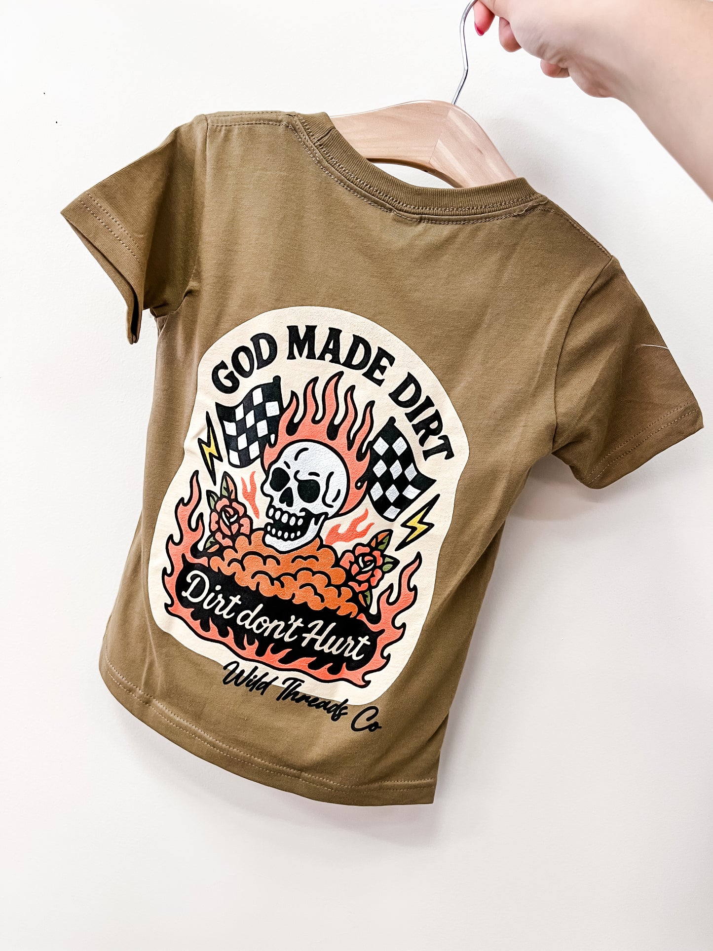 The God Made Dirt T-Shirt by Wild Threads Co is a brown tee displayed on a hanger, featuring a back graphic with a skull, flames, checkered flags, and the phrase "God Made Dirt, Dirt don't Hurt.
