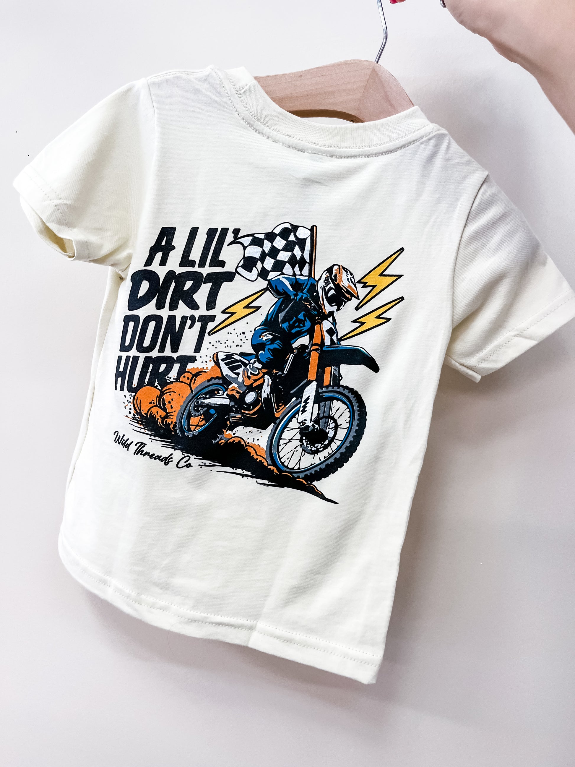The Wild Threads Co A Lil Dirt T-Shirt is a white kids’ tee featuring a dirt bike rider, lightning bolts, and a checkered flag with “A LIL DIRT DON’T HURT” text. Shown held by hand on a hanger.