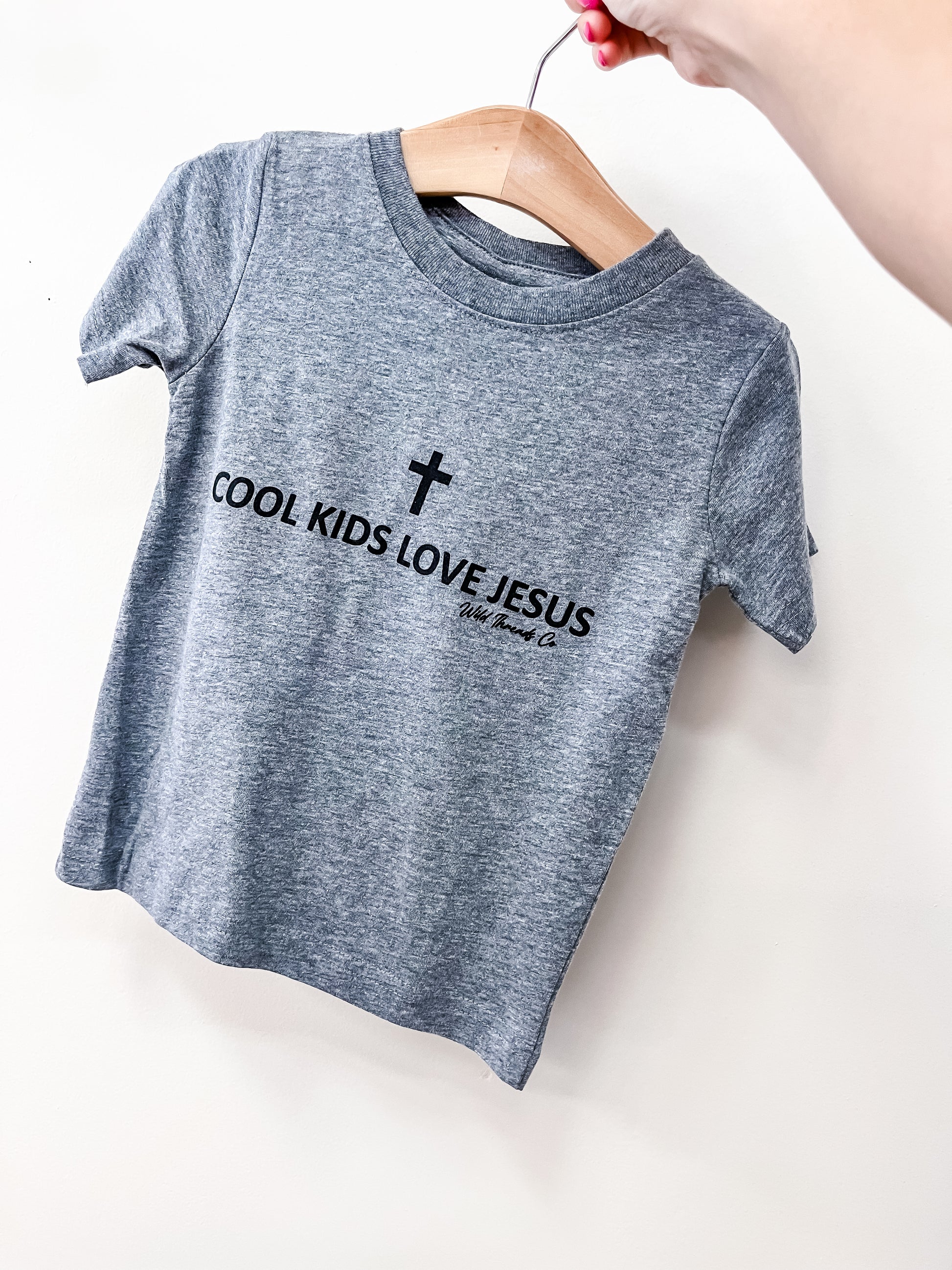 A hand holds a wooden hanger with the Wild Threads Co Faith Kid T-Shirt, a gray children's tee featuring a black cross and "COOL KIDS LOVE JESUS" on the front.