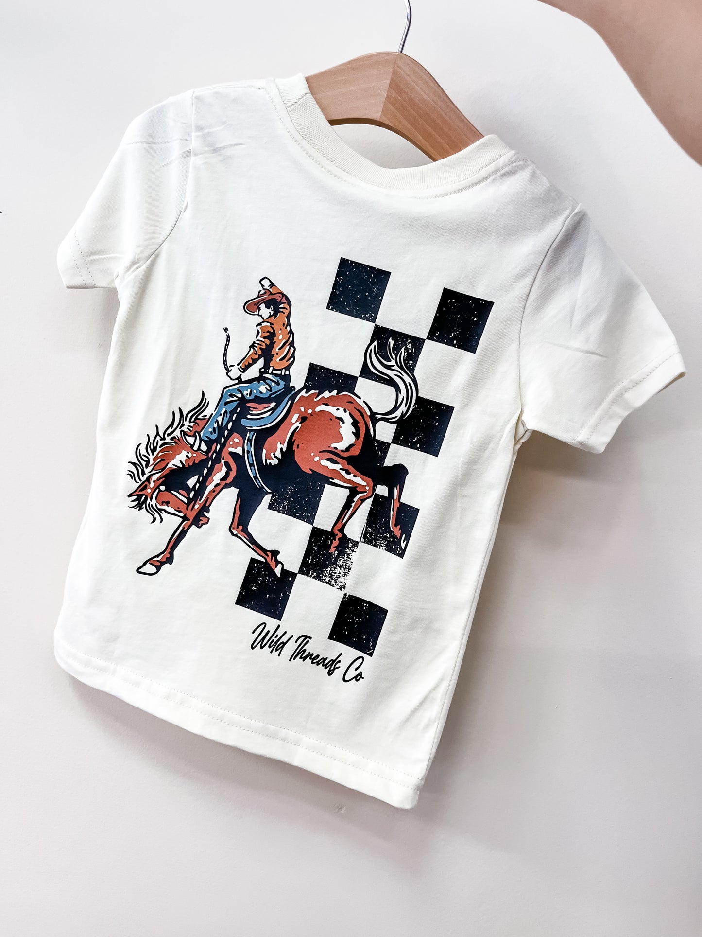 The Little Buckaroo T-Shirt by Wild Threads Co is a white children's tee on a wooden hanger, featuring a rodeo cowboy riding a bucking horse with black-and-white checkerboard background and "Wild Threads Co" text.
