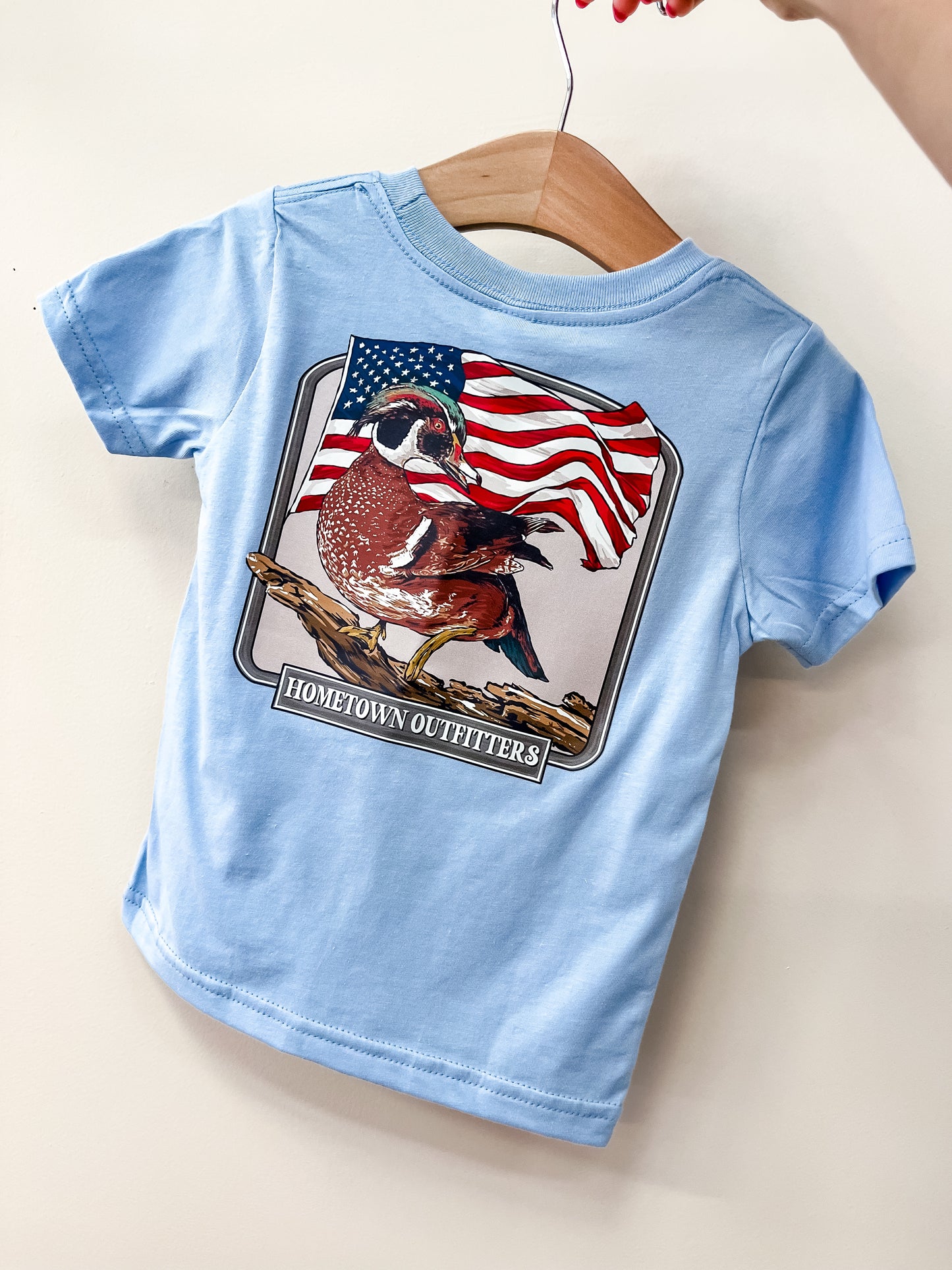 The Little Patriot T-Shirt by Wild Threads Co is a light blue toddler shirt on a wooden hanger, showing a bird on a branch with an American flag behind it and "HOMETOWN OUTFITTERS" below the image.