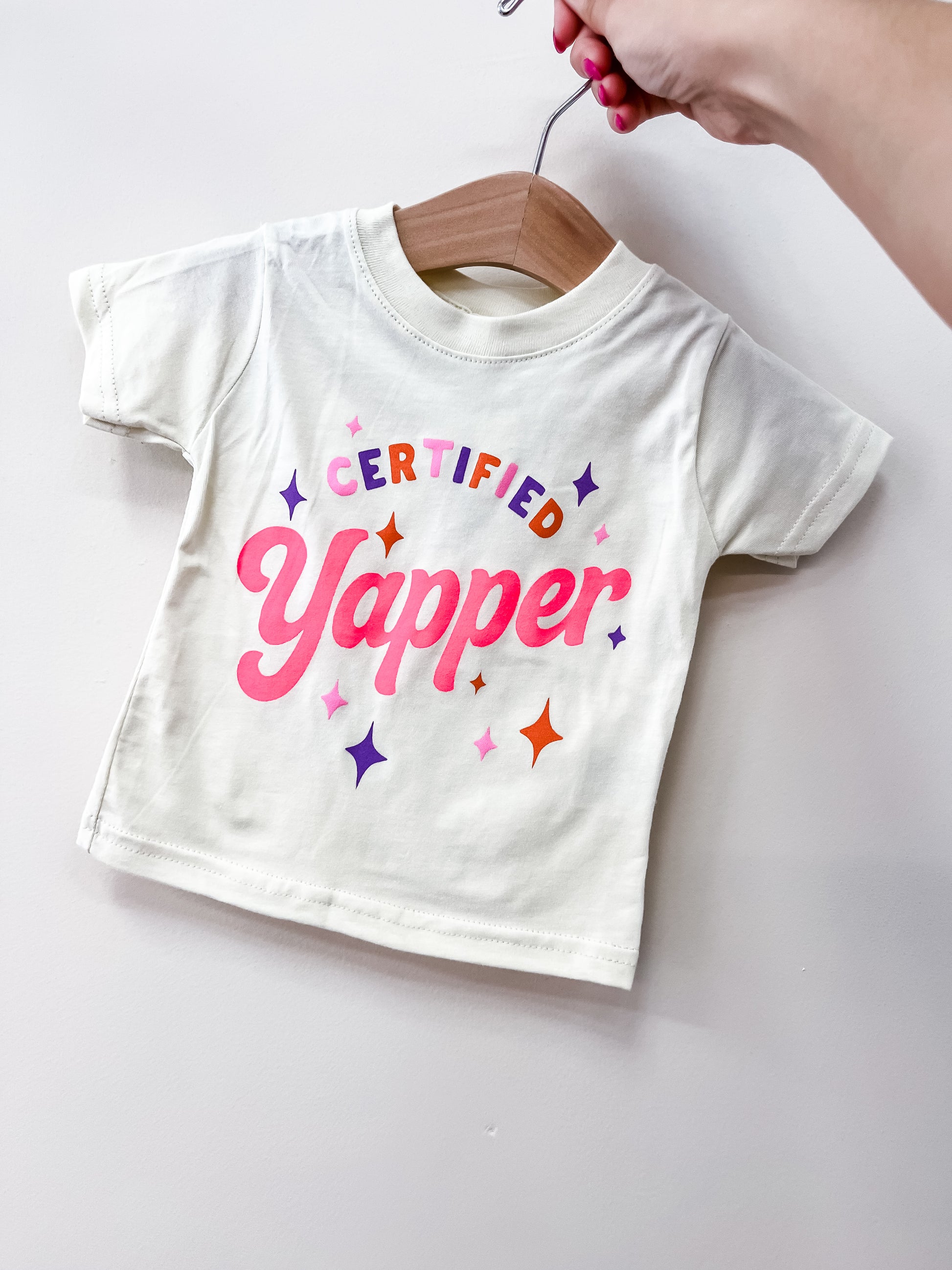 A hand holds up the Wild Threads Co Certified Yapper T-Shirt, a white baby tee on a wooden hanger with colorful text saying "Certified Yapper," surrounded by pink, orange, and purple stars.