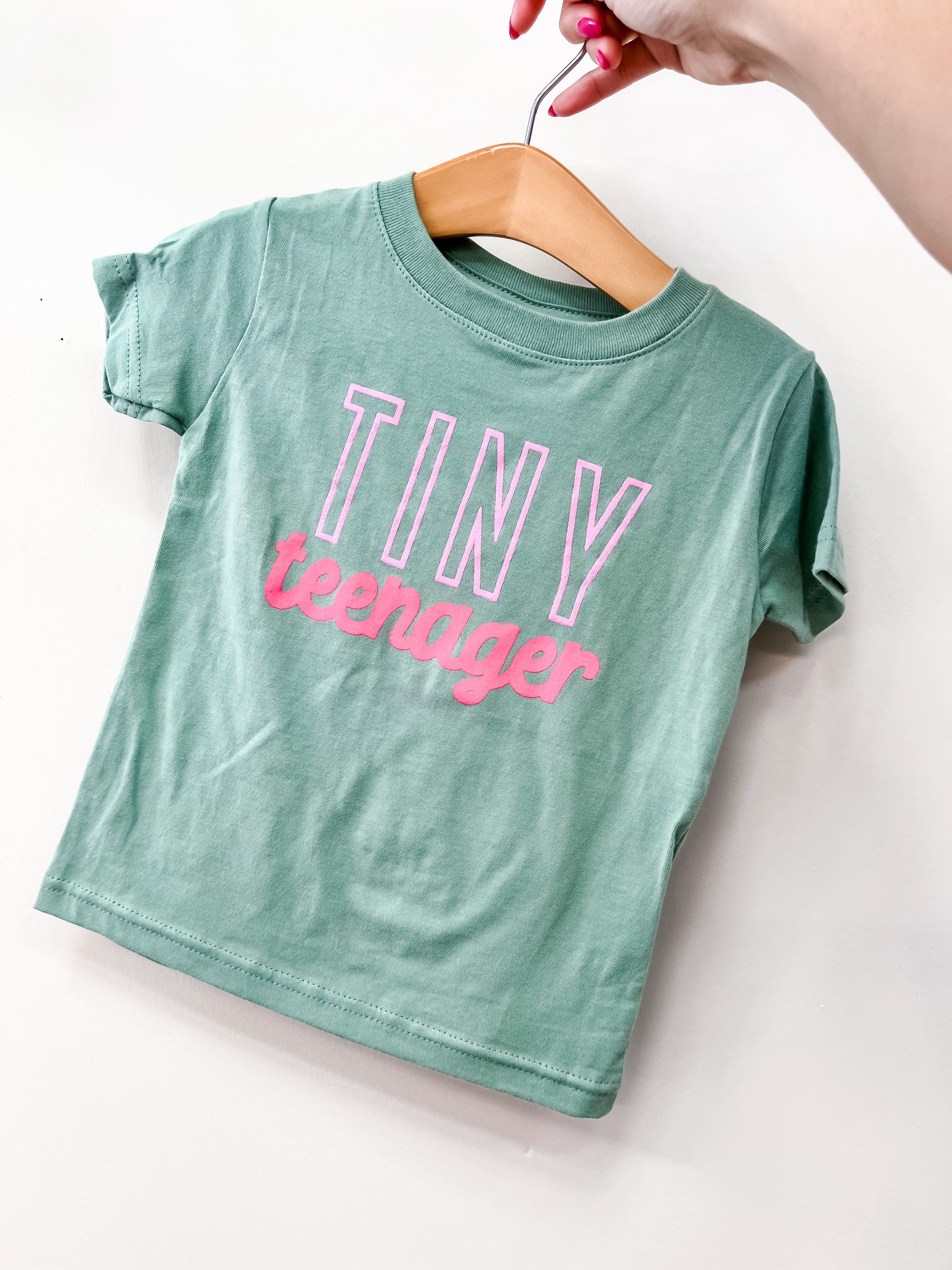 A hand holds a hanger with the Wild Threads Co Tiny Teenager T-Shirt, a small green tee featuring “TINY teenager” in bold white and pink letters.