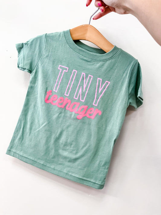 A hand holds a hanger with the Wild Threads Co Tiny Teenager T-Shirt, a small green tee featuring “TINY teenager” in bold white and pink letters.