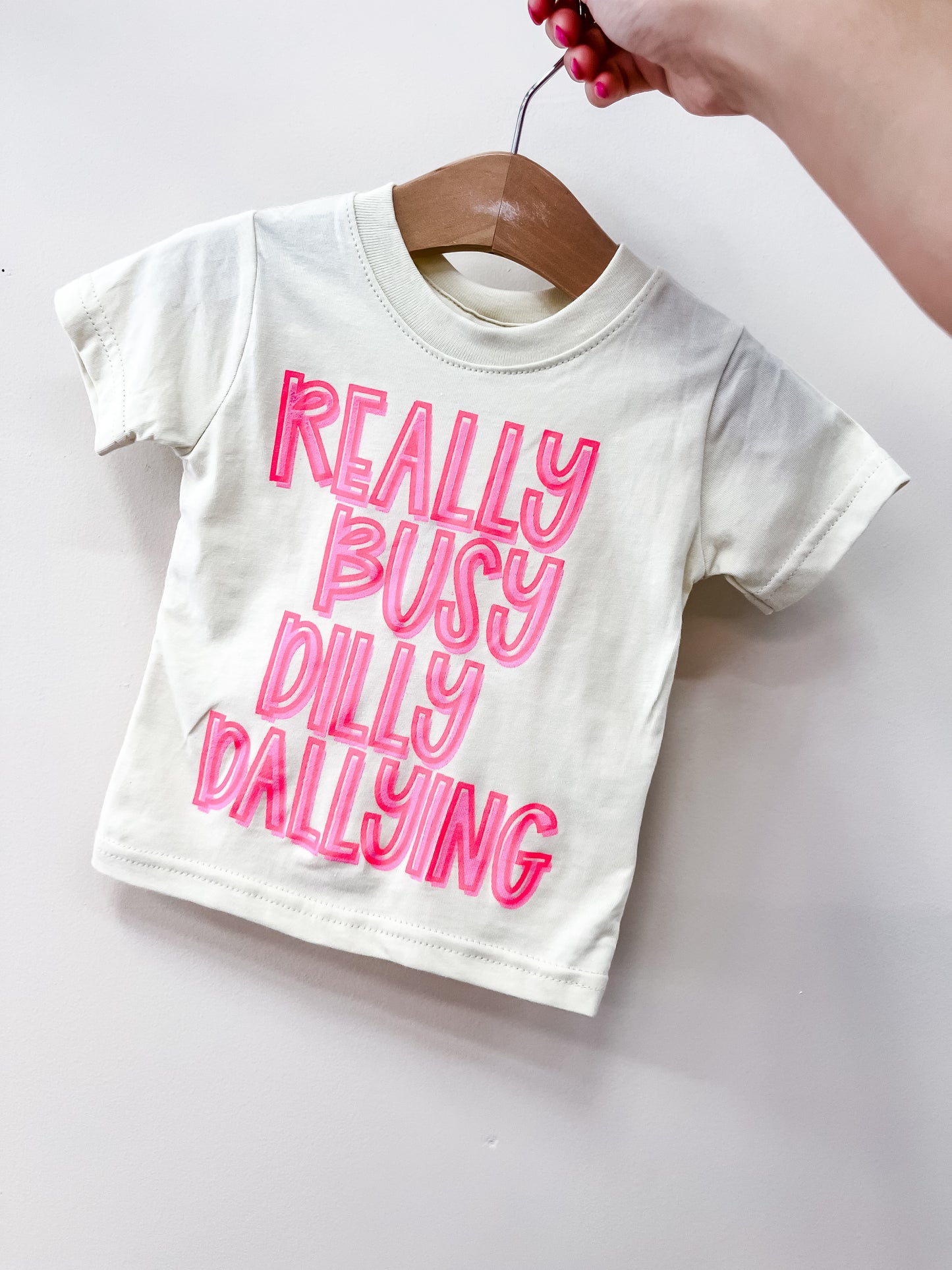 The Dilly Dally T-Shirt by Wild Threads Co is a cream-colored toddler tee with bold, bright pink "REALLY BUSY DILLY DALLYING" text, shown on a wooden hanger held by someone with pink nails.