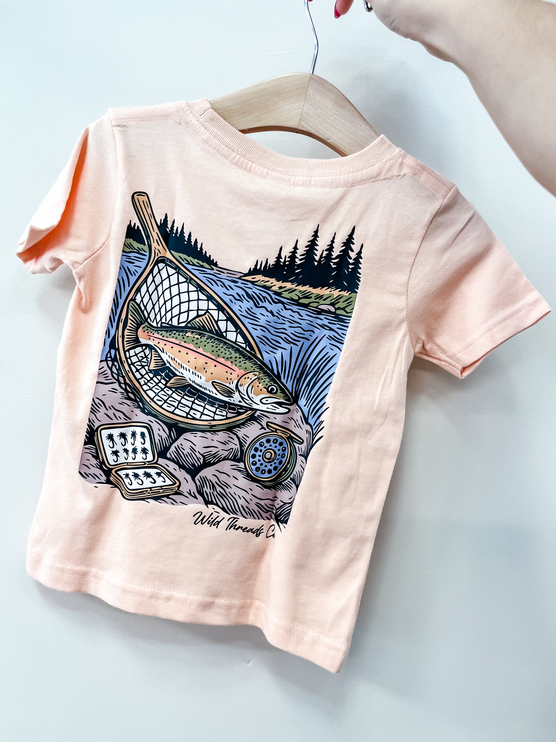 The River Catch T-Shirt by Wild Threads Co is a peach toddler tee on a hanger, featuring a trout in a fishing net by a river, with trees and a box of fishing flies in the background.