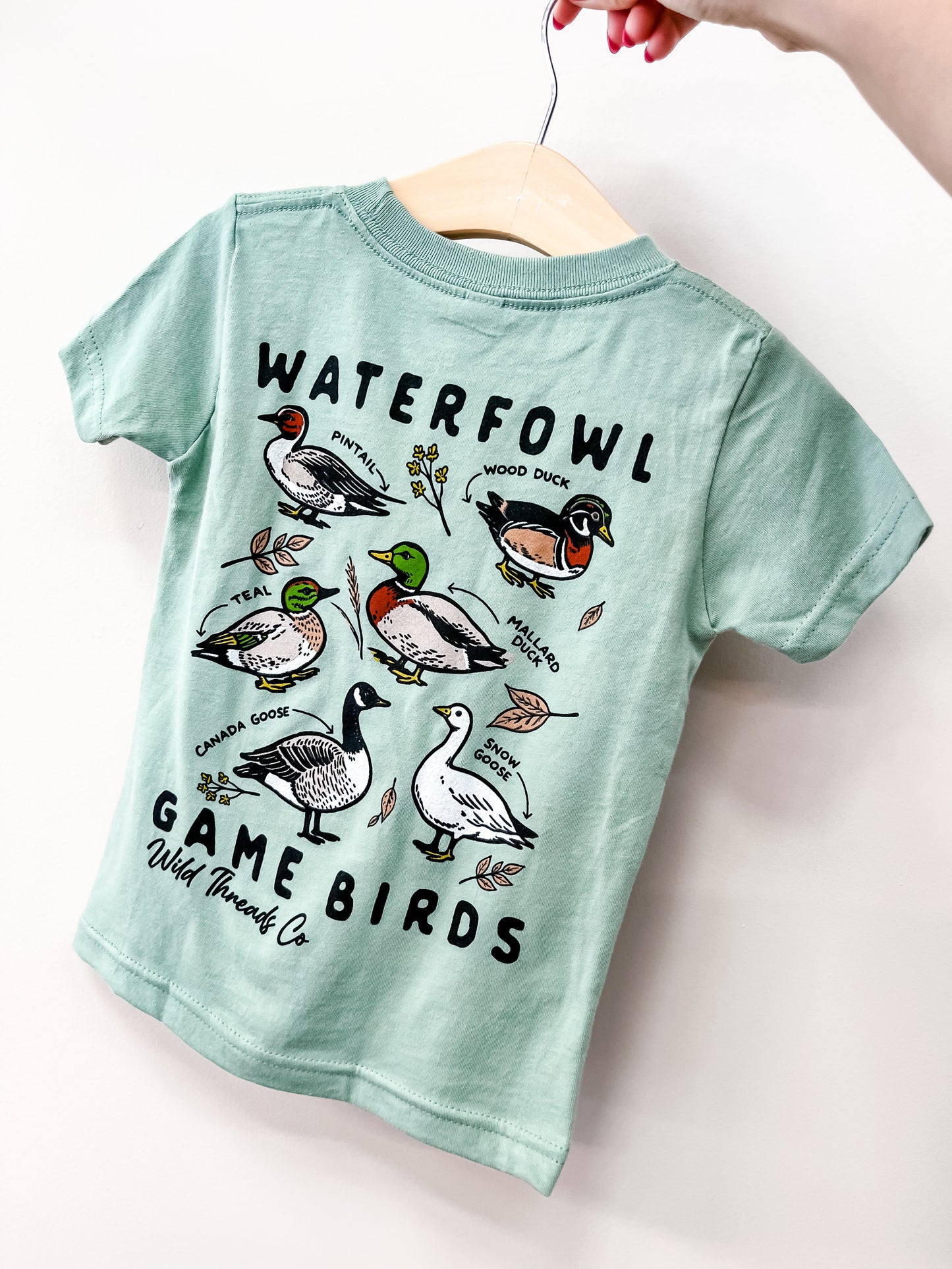 The Little Waterfowl T-Shirt by Wild Threads Co is a light green kids' tee on a hanger, featuring illustrated teal, pintail, mallard, wood duck, Canada goose, and snow goose with "Waterfowl Game Birds" text.