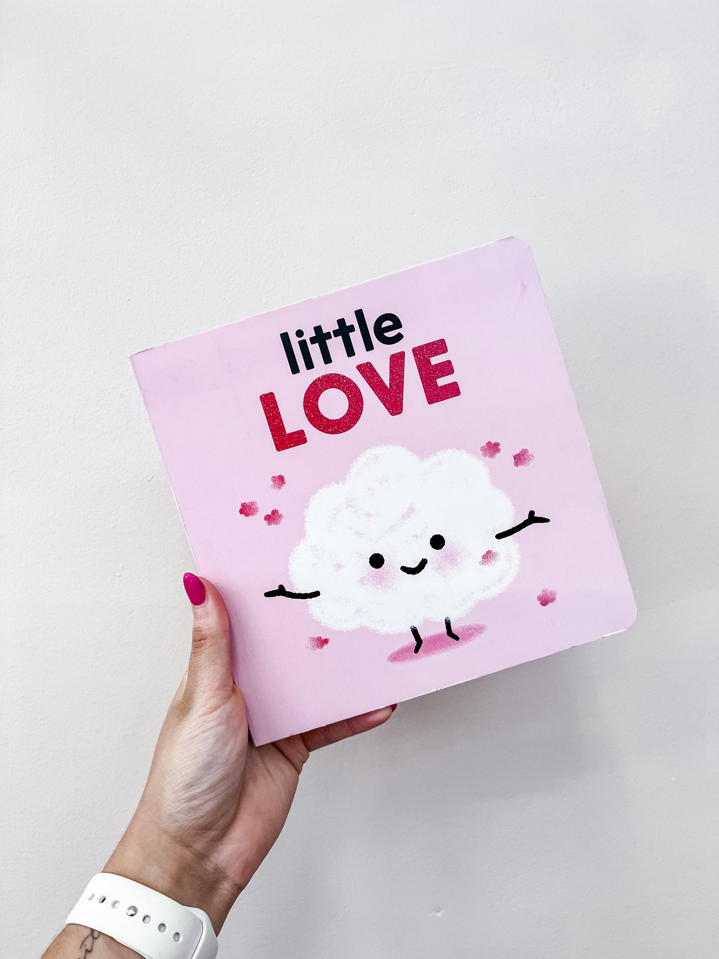 A hand with pink-painted nails holds the Little Love Book by Tiny Tot Bundles, featuring a smiling fluffy white cloud with stick arms and legs on the cover, surrounded by small pink hearts.